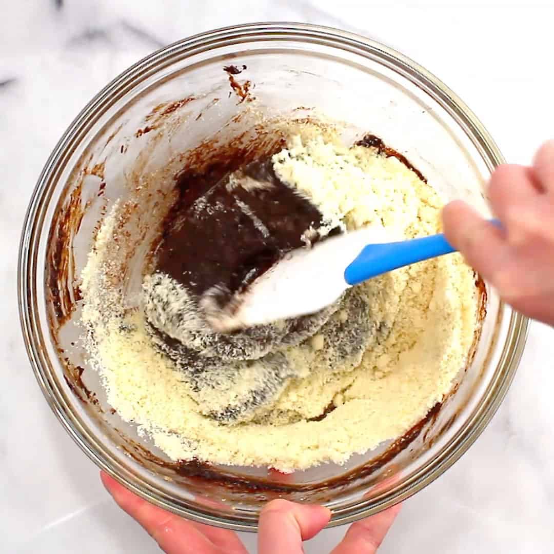 hand holding rubber spatula folding flour into wet ingredient brownie mixture
