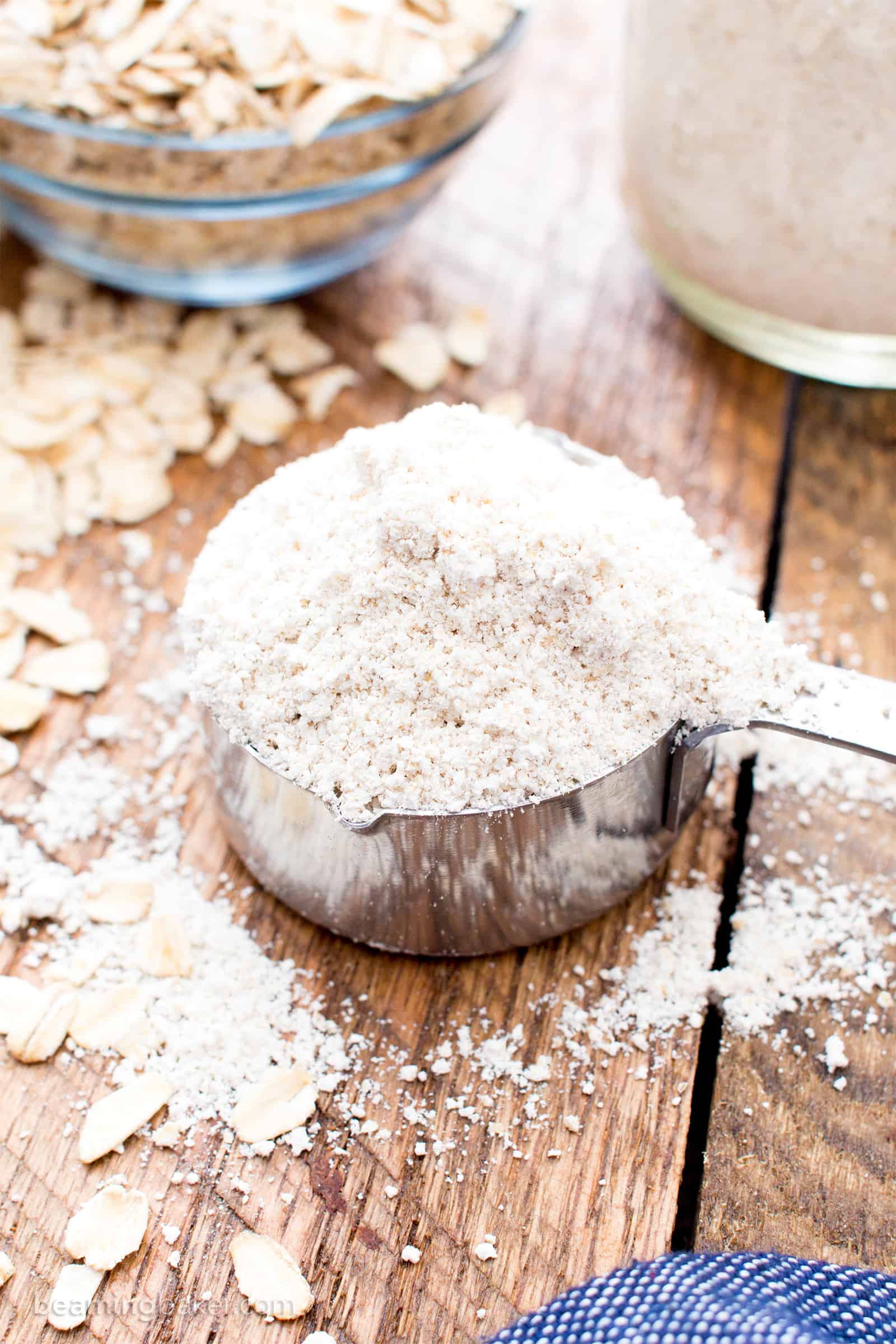 Measuring cup filled with homemade gluten free oat flour sitting on a kitchen table, with loose oats and oat flour scattered around it