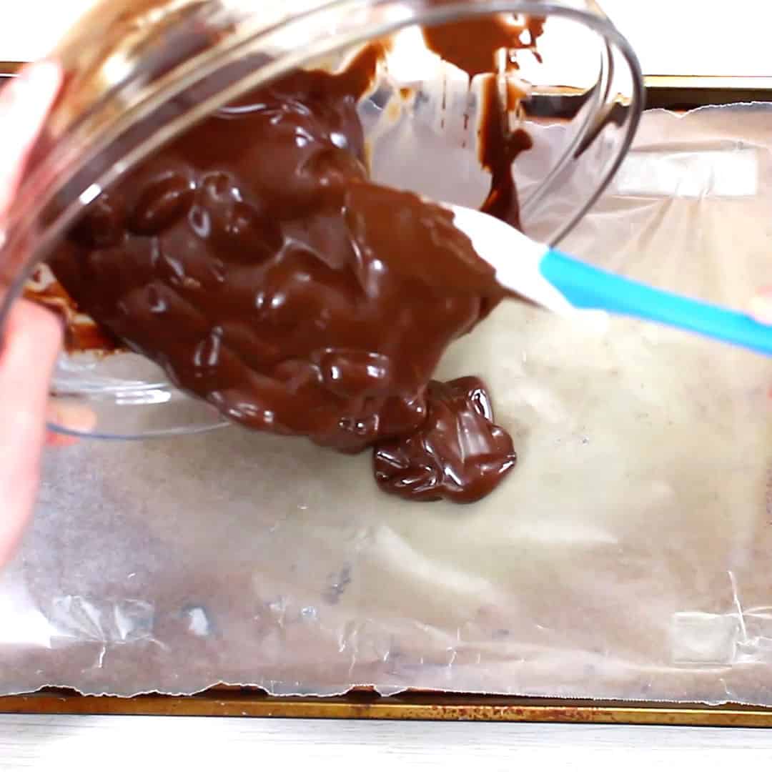 hand holding a bowl over baking sheet pouring melted chocolate almond bark mixture