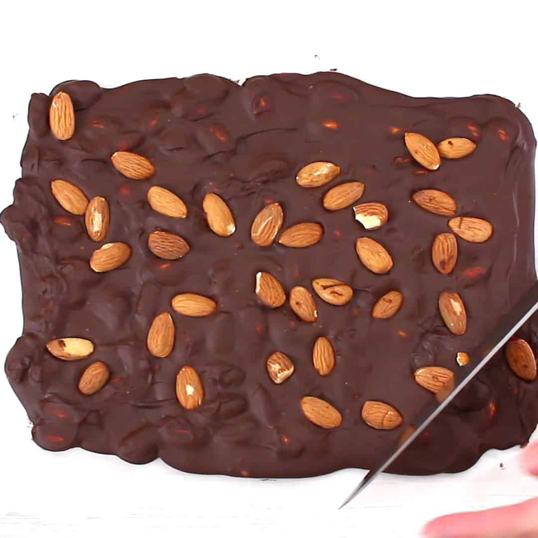 hand holding knife to cut sheet of hardened chocolate almond mixture into almond bark pieces
