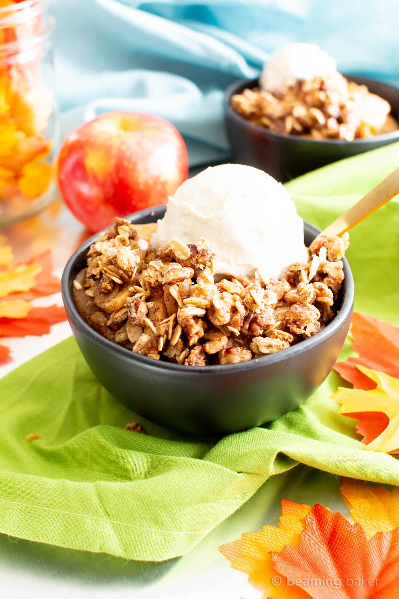 Black bowl of vegan gluten free apple crisp with vegan vanilla ice cream on top, sitting on green cloth with additional bowl and whole apple in the background