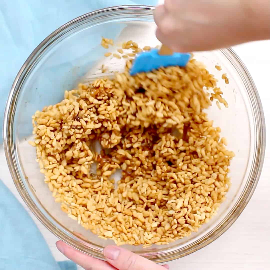 A hand folding and stirring rice crisp cereal into sweetened mixture for rice crispy treats
