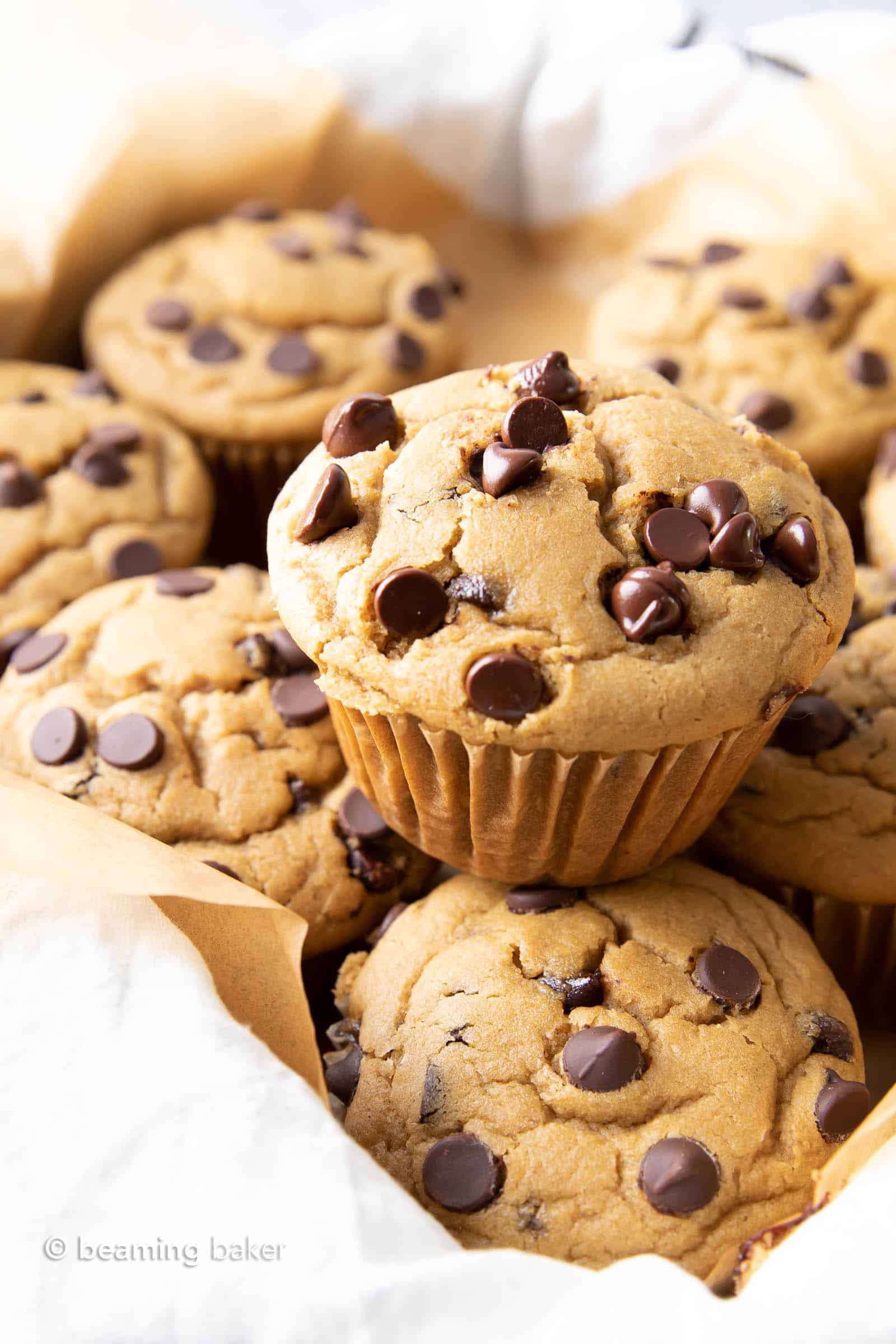 Chocolate chip muffins on parchment paper and cloth in a tray, with a single muffin stacked on top