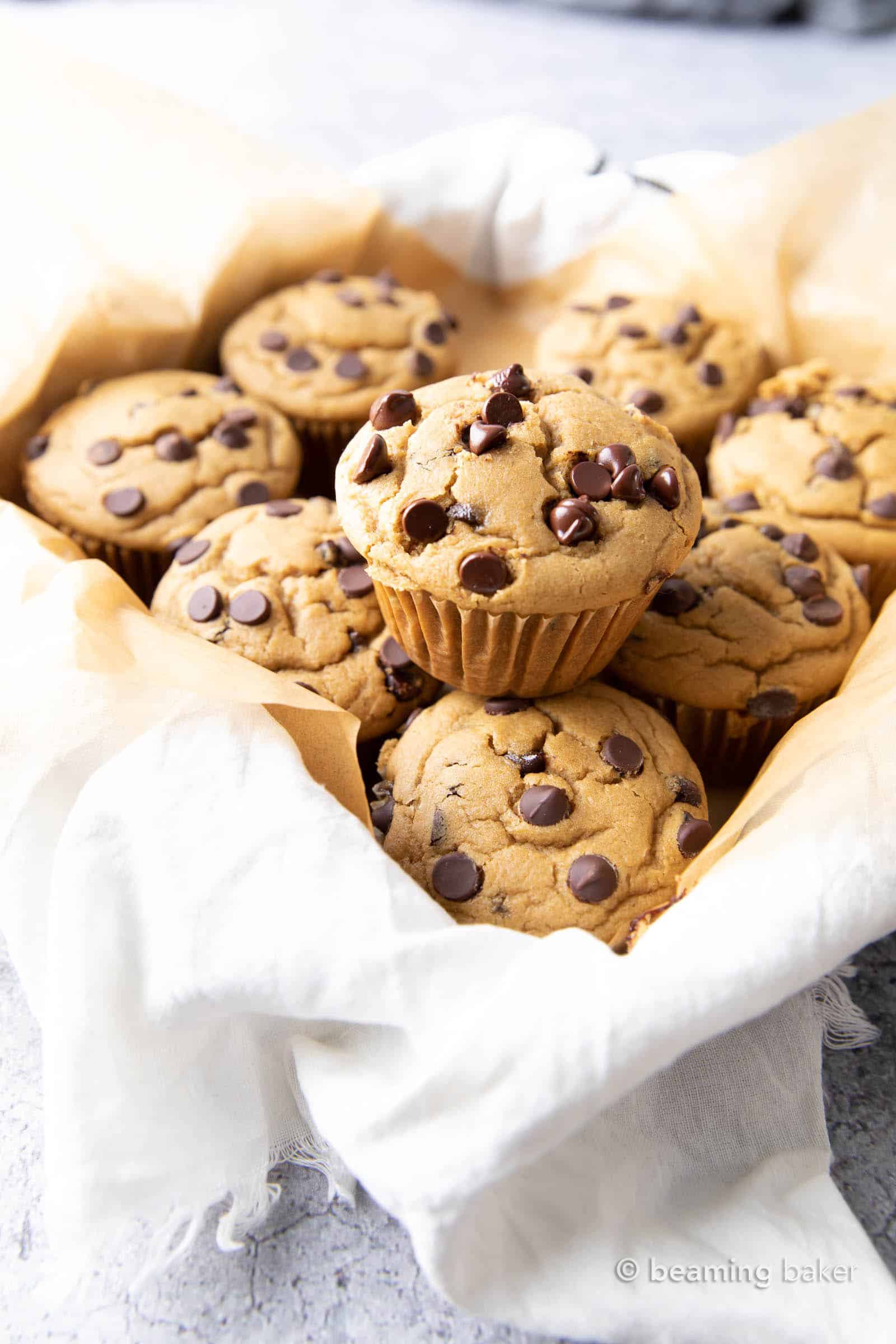 Angled shot of healthy chocolate chip muffins in a tray with parchment paper and cloth, with a single muffin stacked on top of the rest