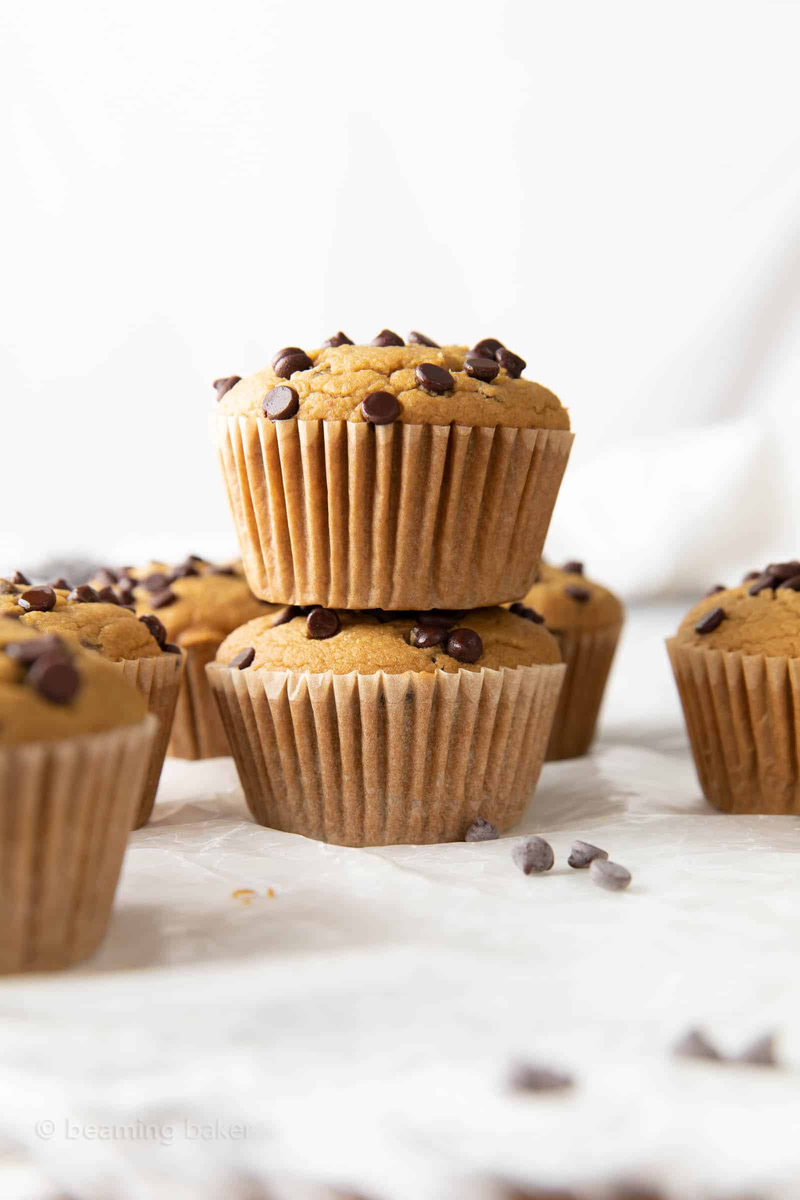 a stack of healthy chocolate chip muffins cooling on parchment paper with more chocolate chips muffins