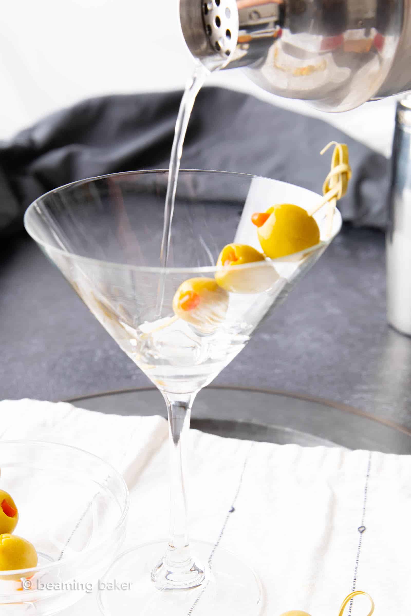 Closeup shot of pouring dirty martini liquid into a chilled martini glass over olive skewer