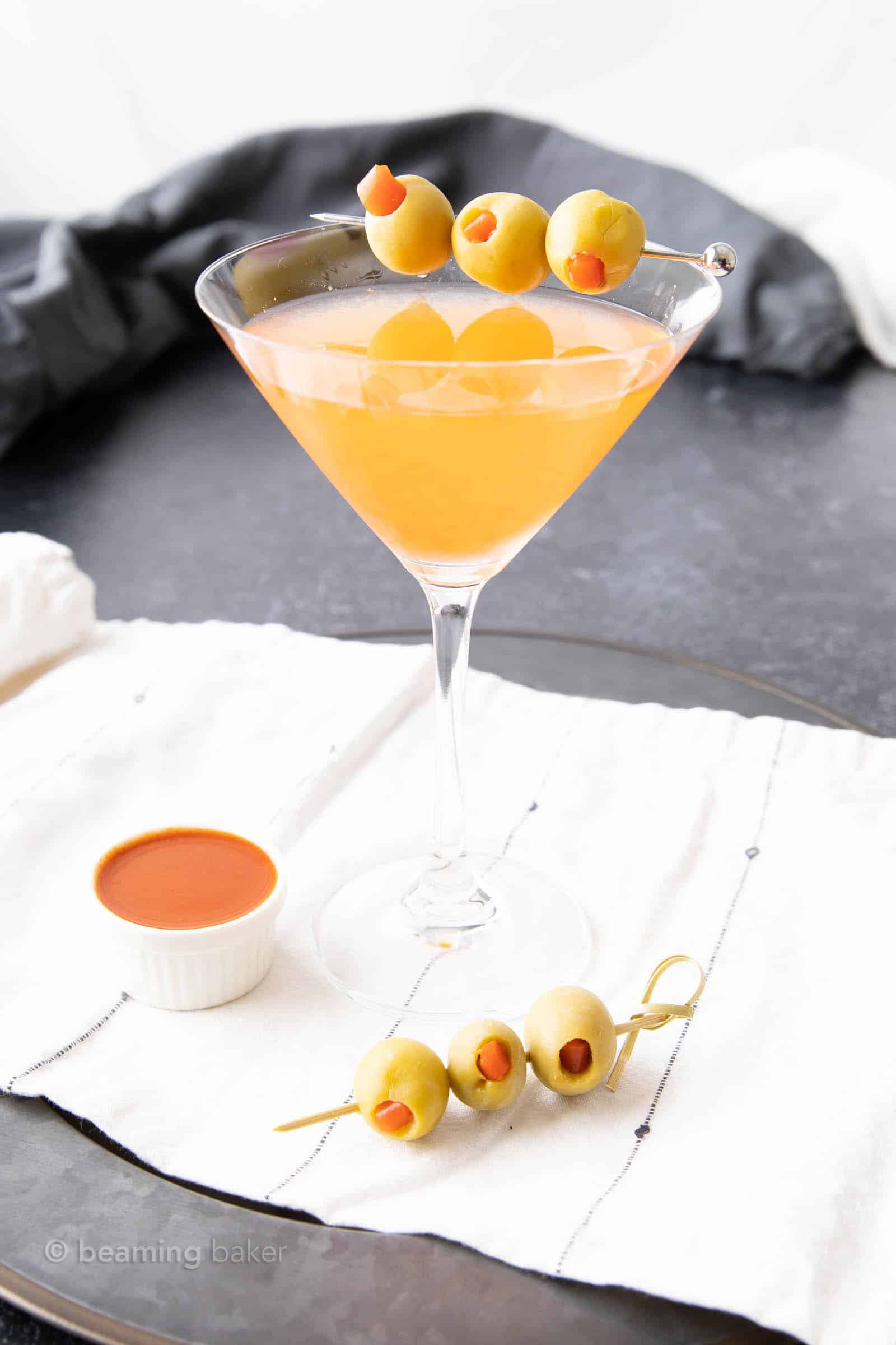 large martini glass with hot and dirty martini recipe in it and olive skewer on napkin nearby