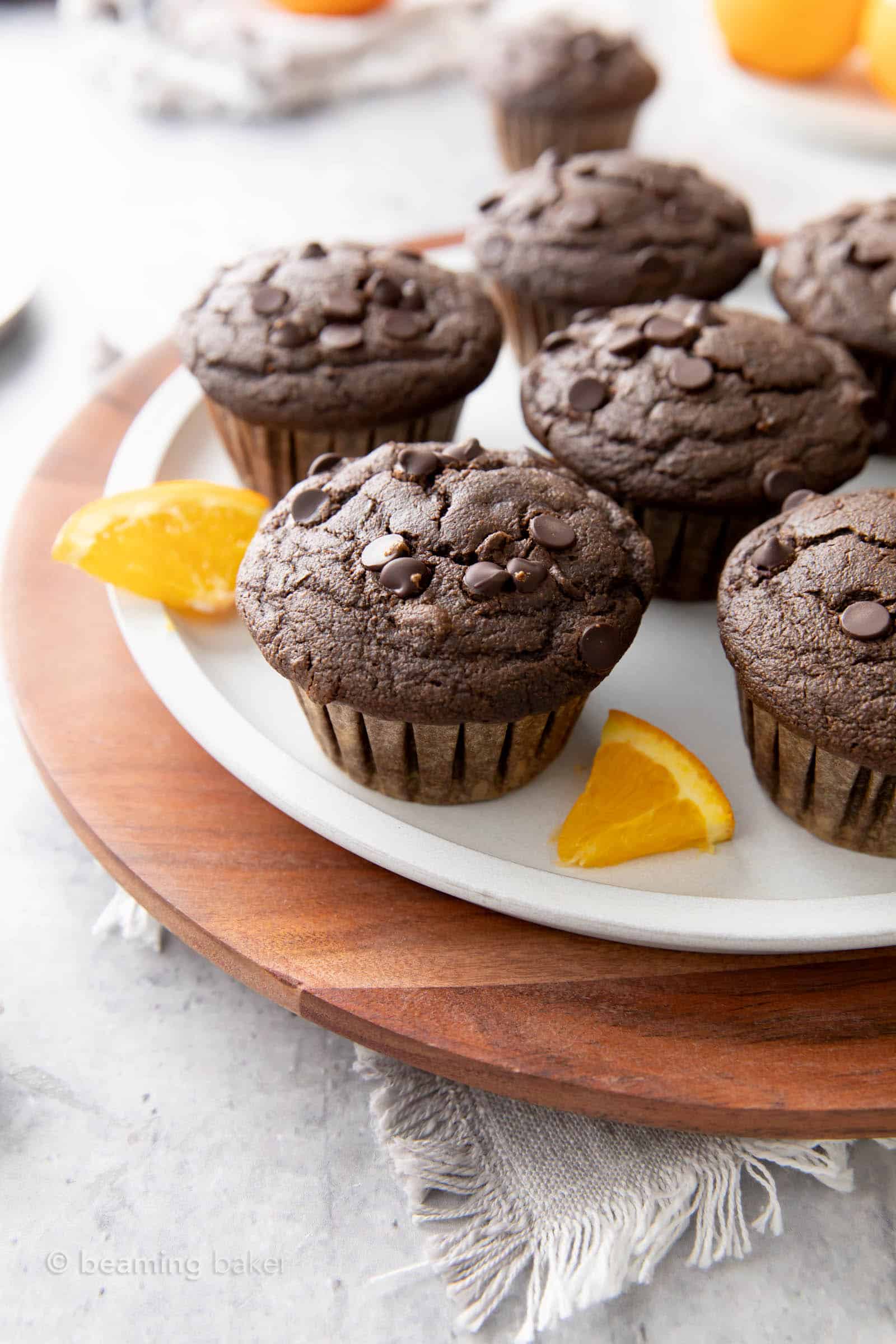 Many vegan chocolate orange muffins served on a plate and serving tray with a kitchen towel and fresh oranges