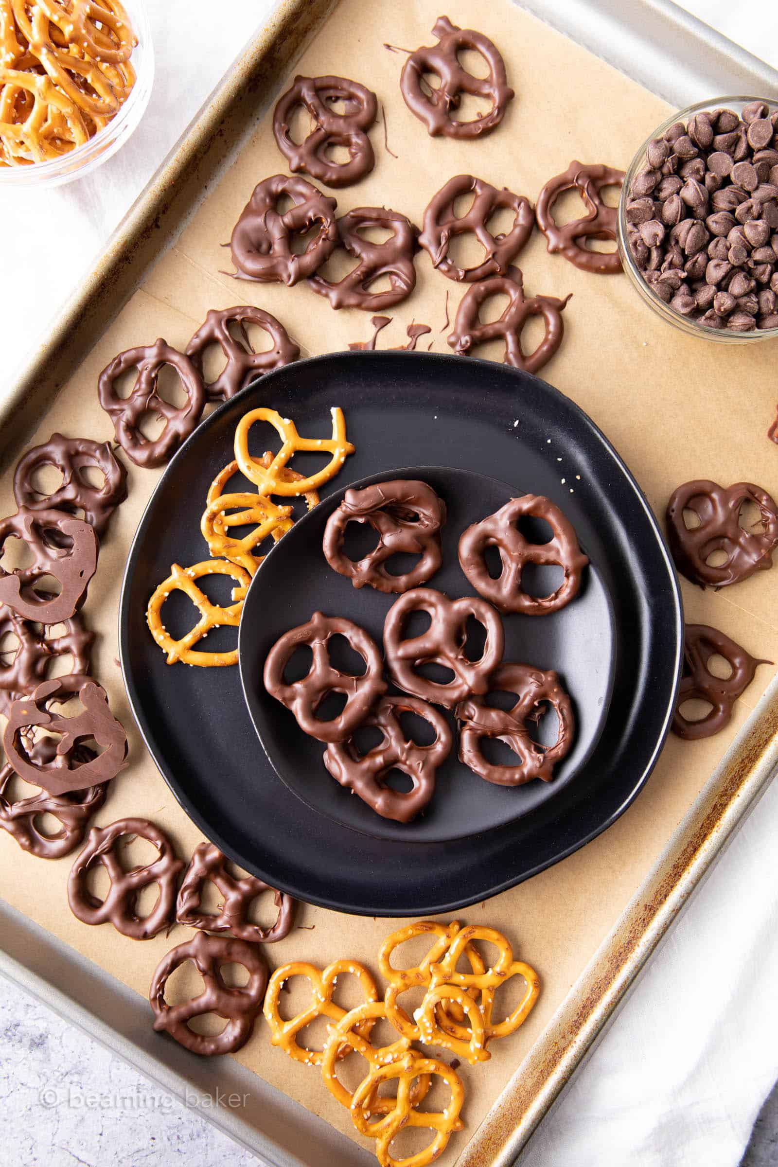 Plate of vegan chocolate covered pretzels on a sheet with more chocolate covered pretzels chocolate and regular pretzels