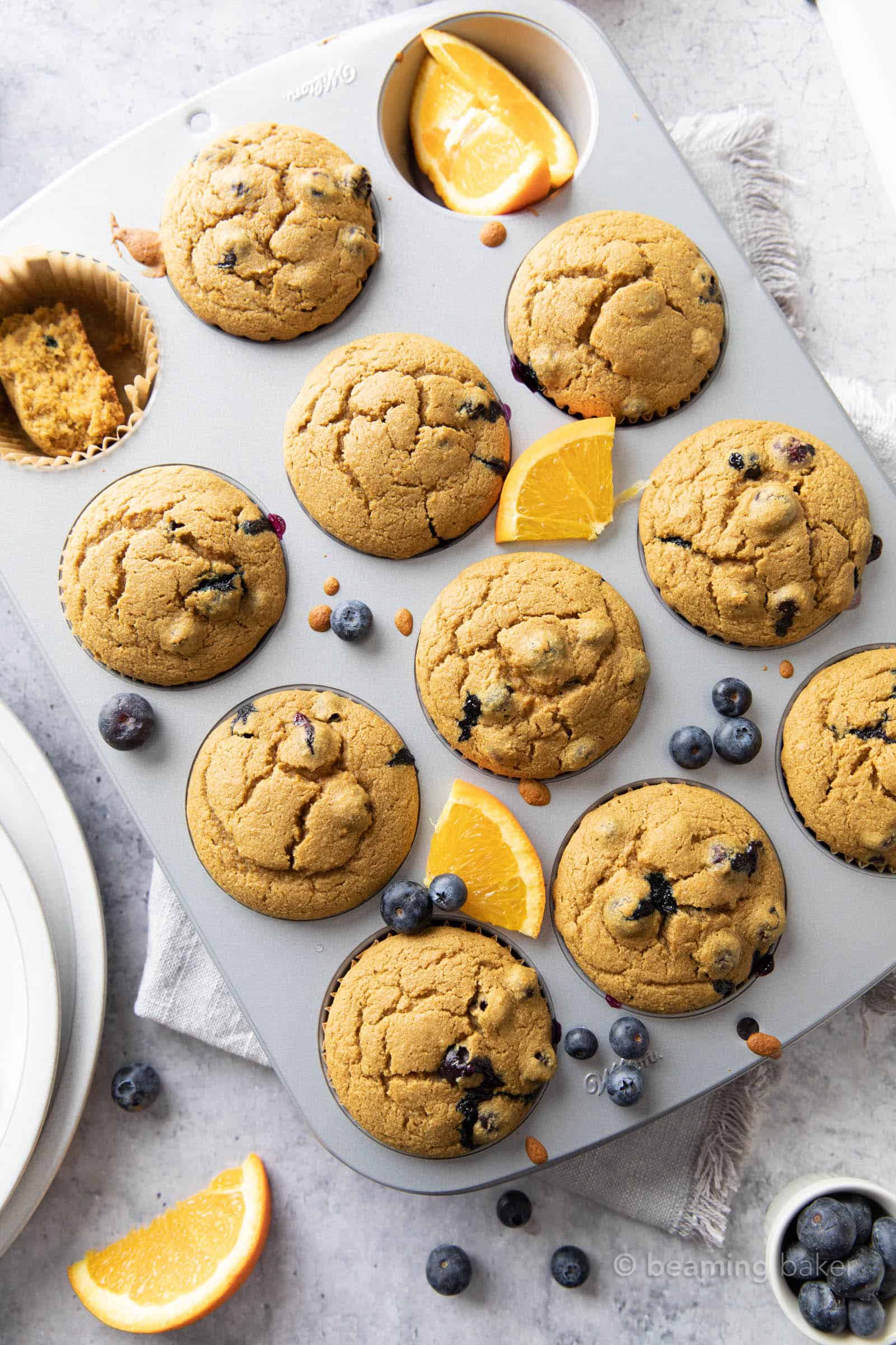 Fresh baked blueberry orange muffins in a metal muffin pan with blueberries and oranges