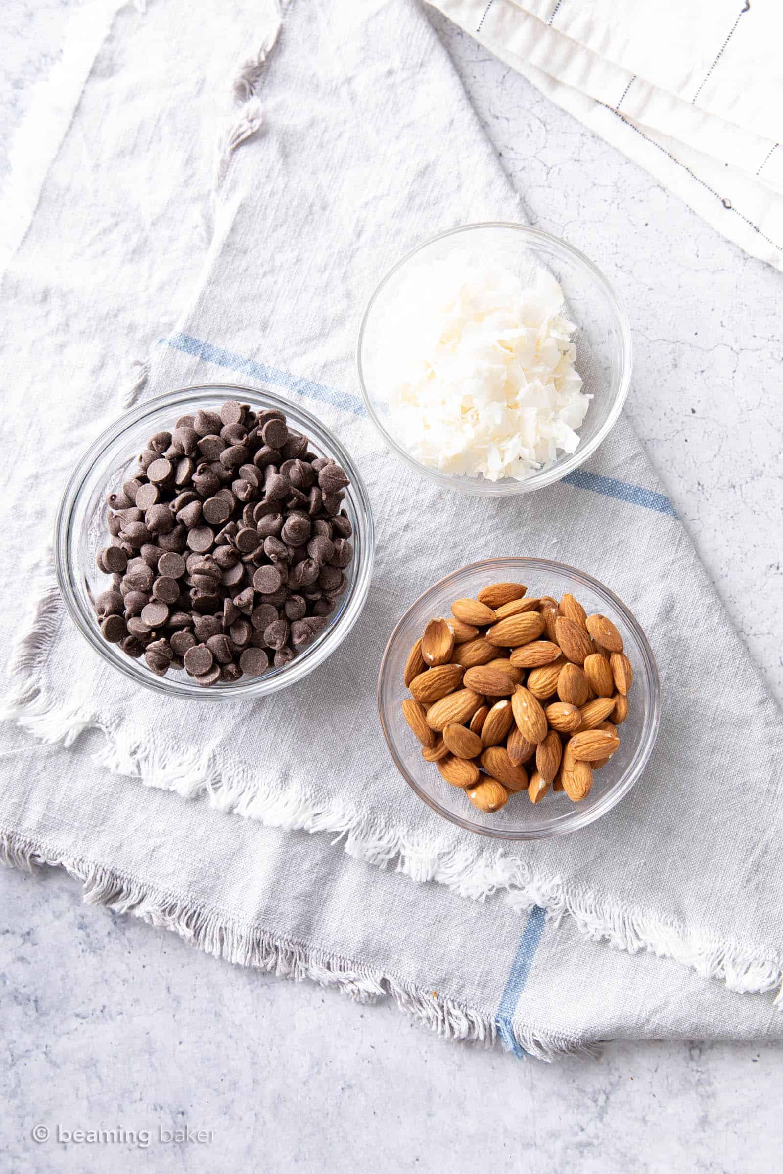 Three mis en place glass bowls filled with dark chocolate chip, roasted almonds, and coconut chips