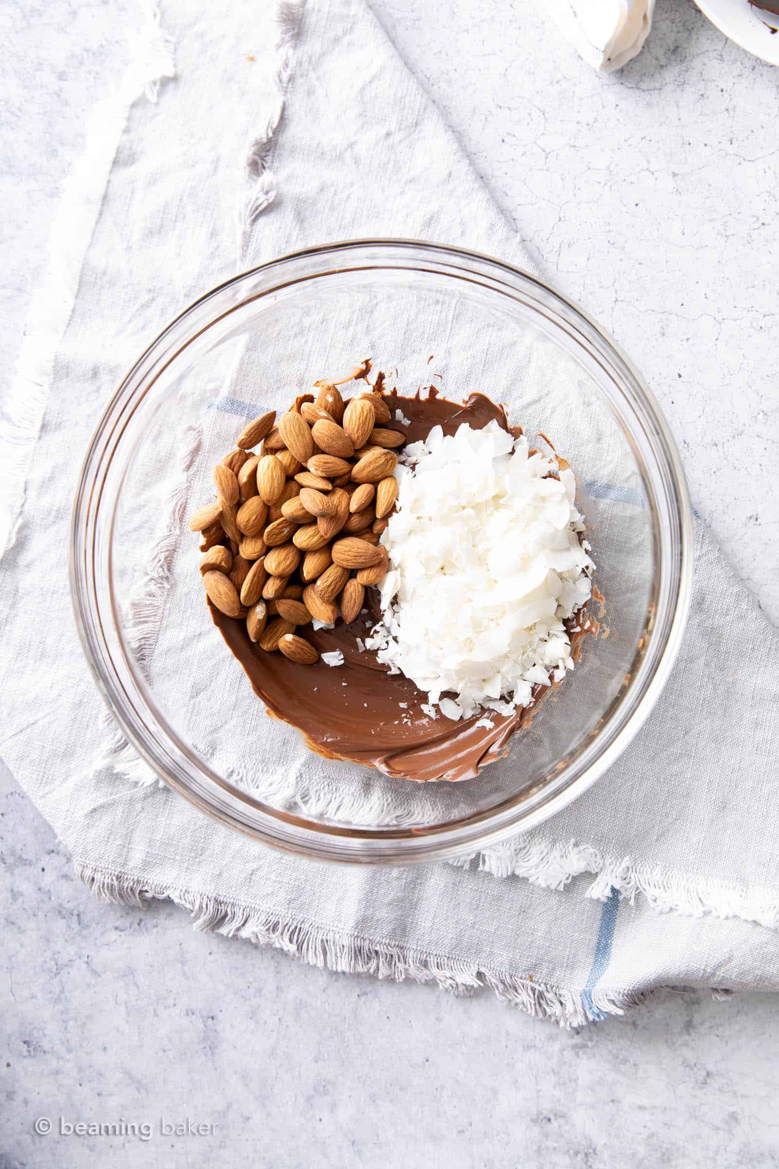 Roasted almonds and coconut chips sitting on top of melted dark chocolate in a glass bowl