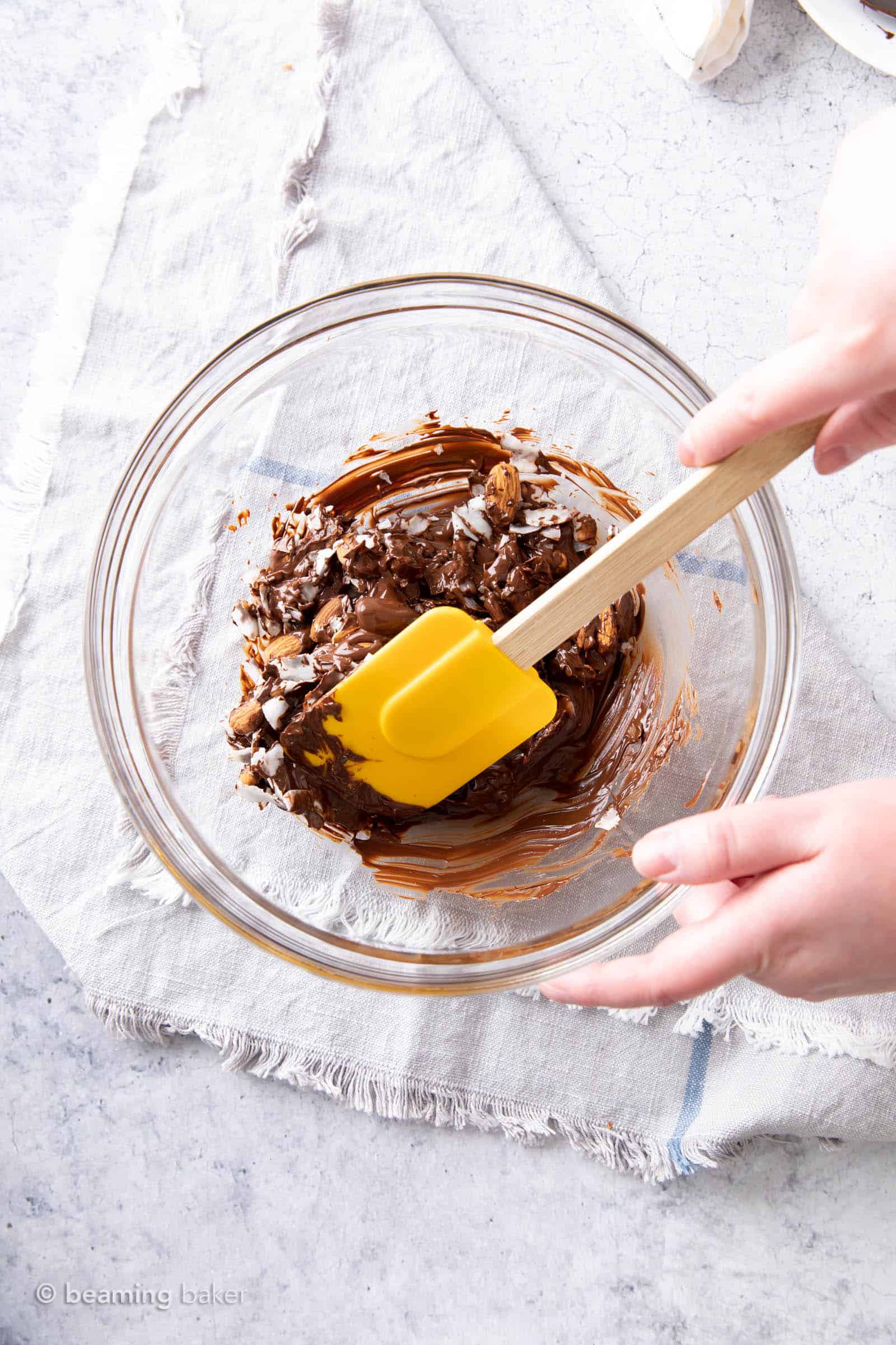 One hand holding a bowl while another hand holds a spatula to fold coconut and almonds into dark chocolate mixture