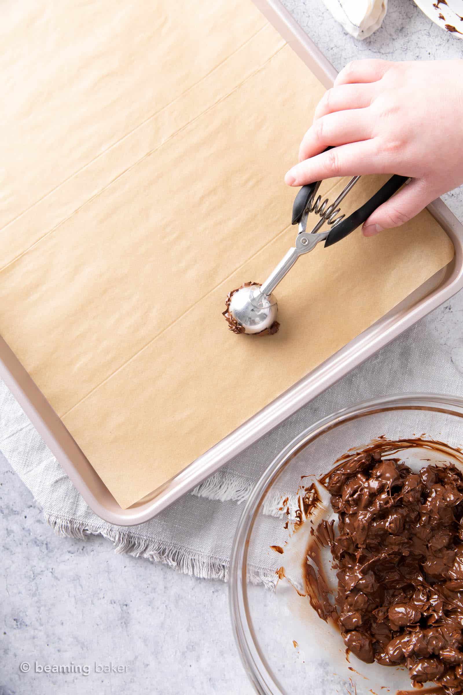One hand holding a food scoop, dropping dark chocolate almond coconut mixture onto a lined baking sheet