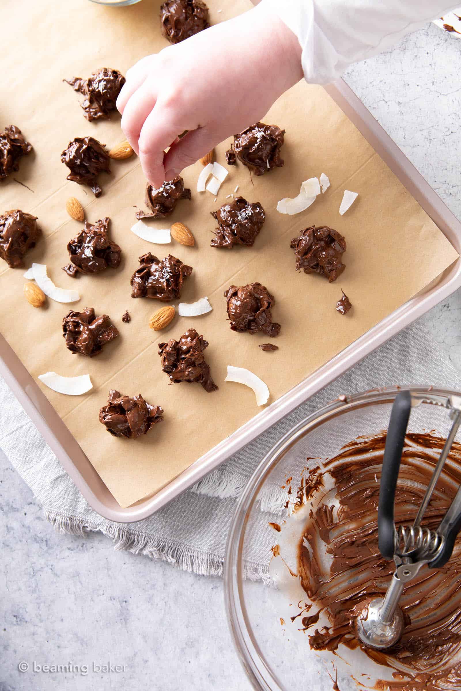 A hand sprinkling chopped almonds and shredded coconut onto melted dark chocolate almond coconut clusters with a bowl of melted chocolate nearby