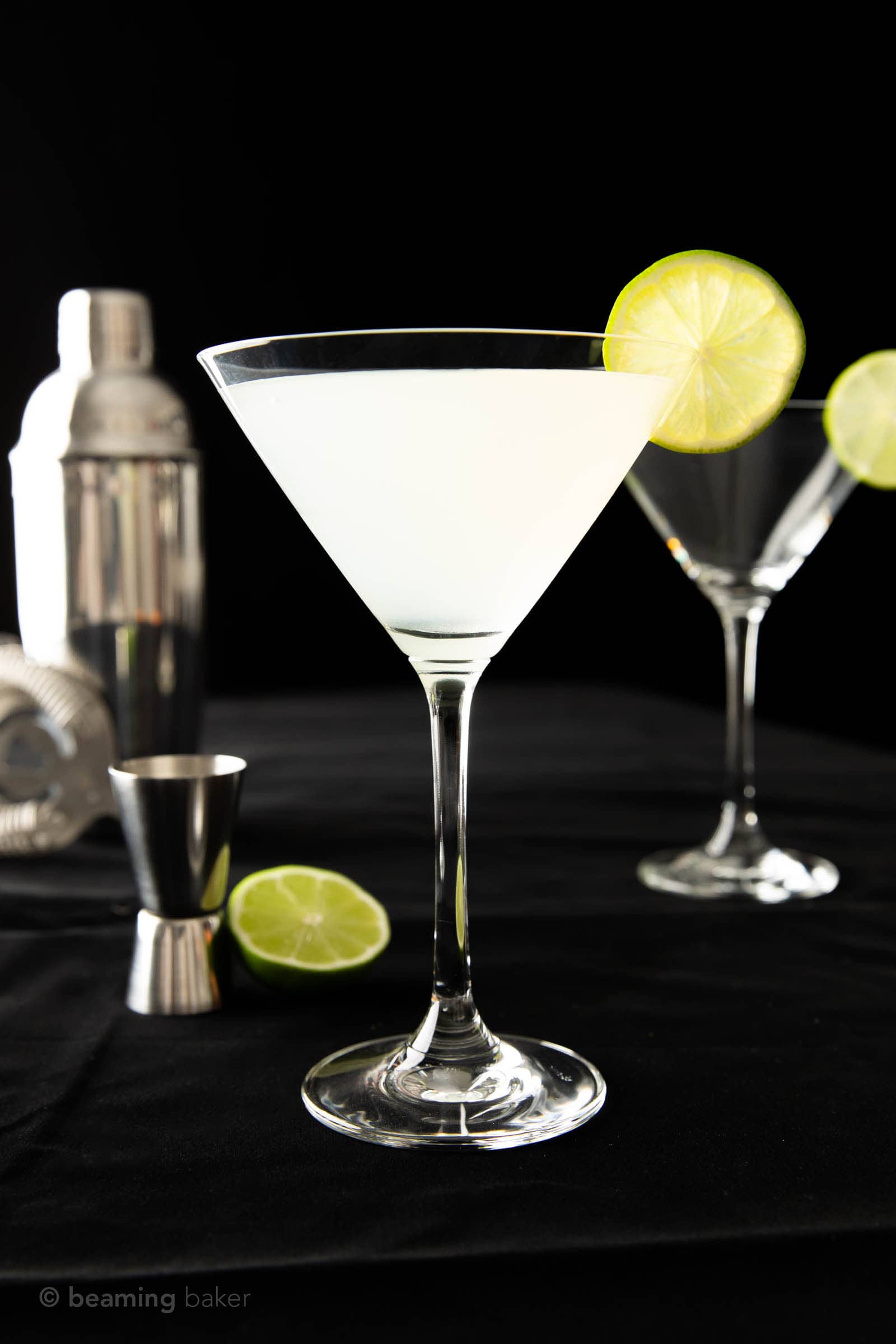 A gimlet recipe in one martini glass with a cocktail shaker jigger and strainer with lime wedges