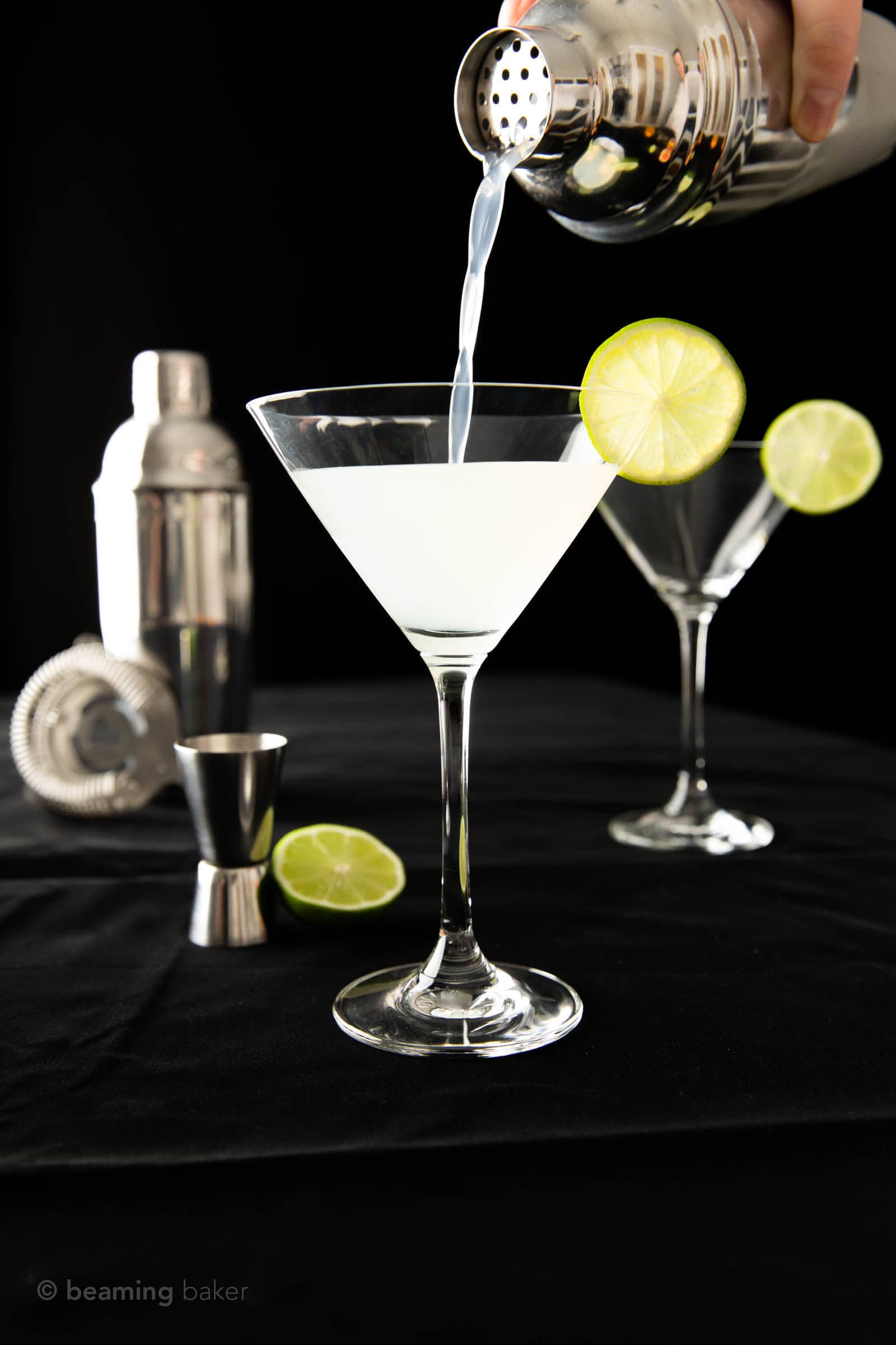 The last pour of gimlet cocktail from a cocktail shaker into one of two martini glasses