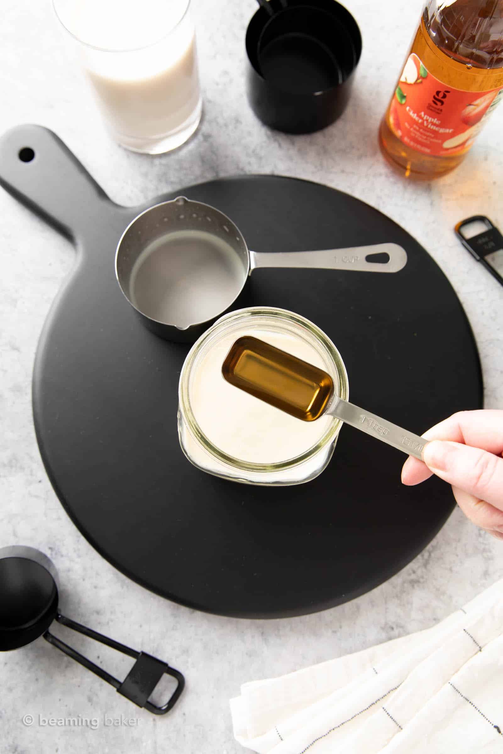 A hand holding one tablespoon of vinegar above a glass jar filled with one cup of soy milk