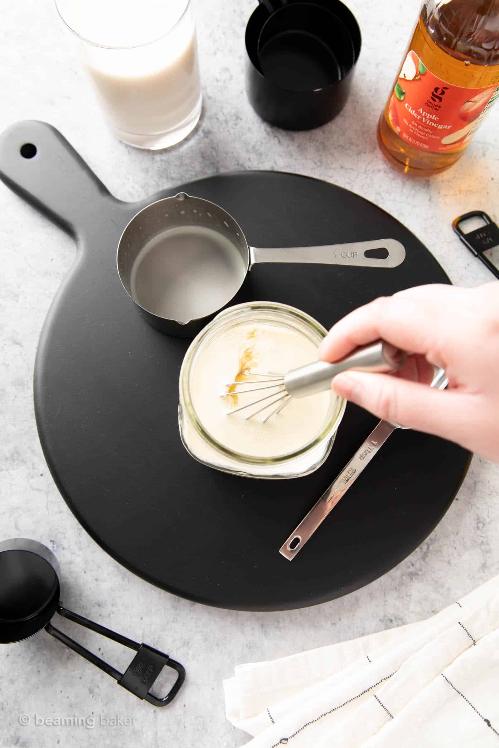 a hand holding a small whisk that is whisking apple cider vinegar with soy milk to make the vegan substitute for buttermilk