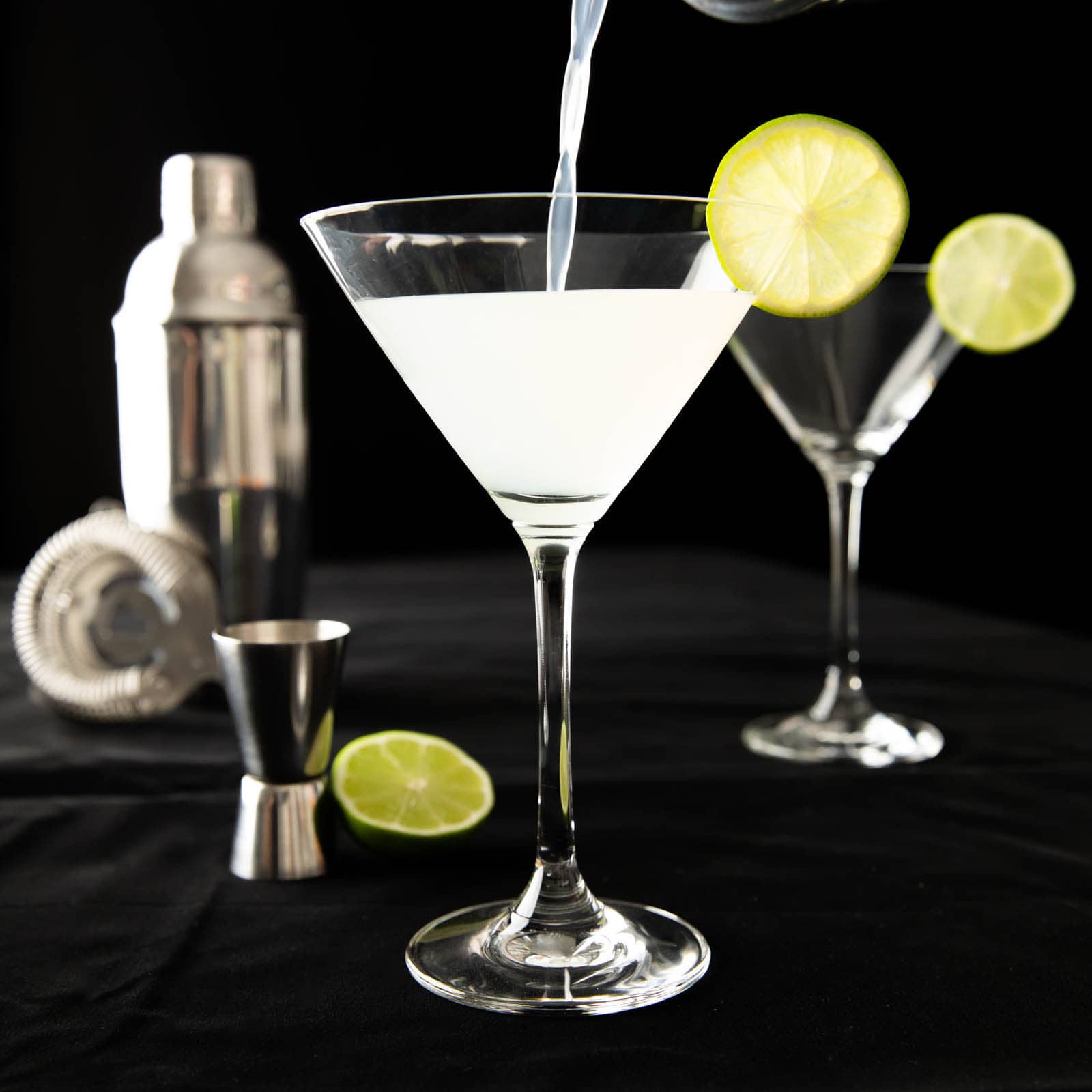 Pouring Gimlet cocktail mixture from a cocktail shaker into a glass with a lime wedge garnish