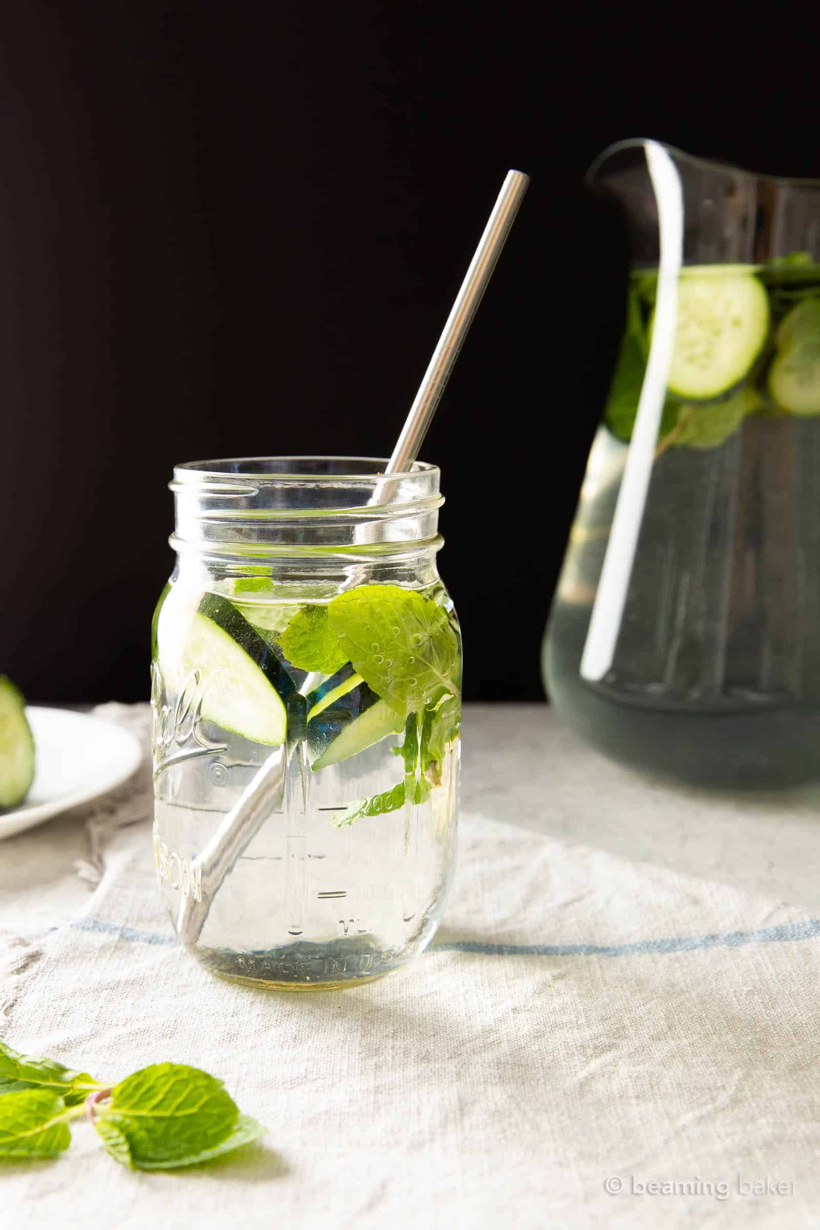 Glass of cucumber mint water with a metal straw inside, and pitcher in the background