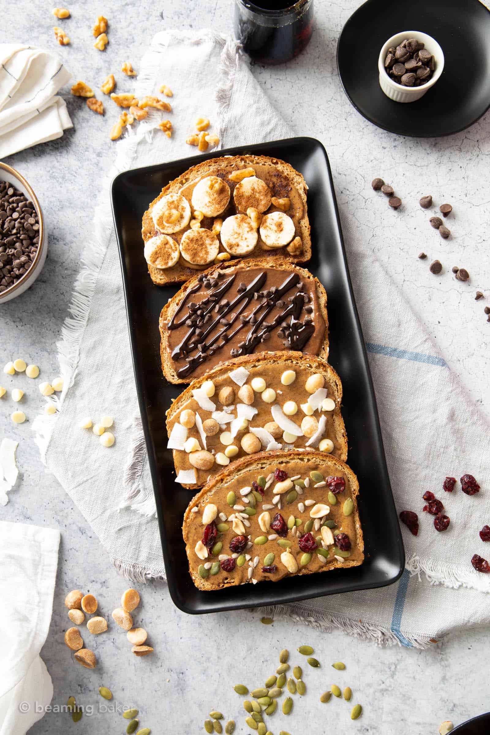 kitchen table covered in platter of almond butter toast recipes with nuts and other toppings in stoneware bowls