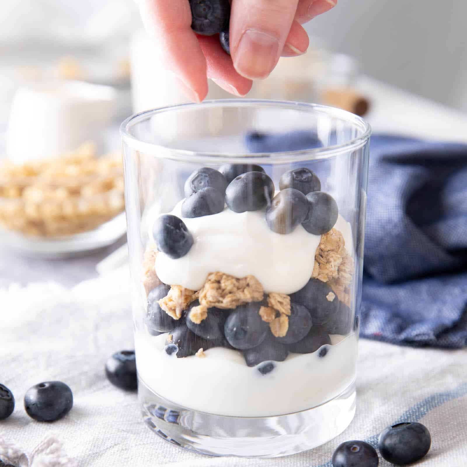 Hand dropping fresh blueberries over layers of yogurt, granola, and blueberries into a glass