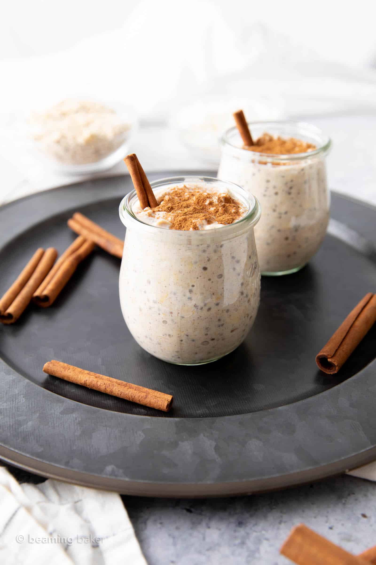 Cinnamon sticks and jars of overnight oats sitting on a gray serving tray with a white cloth