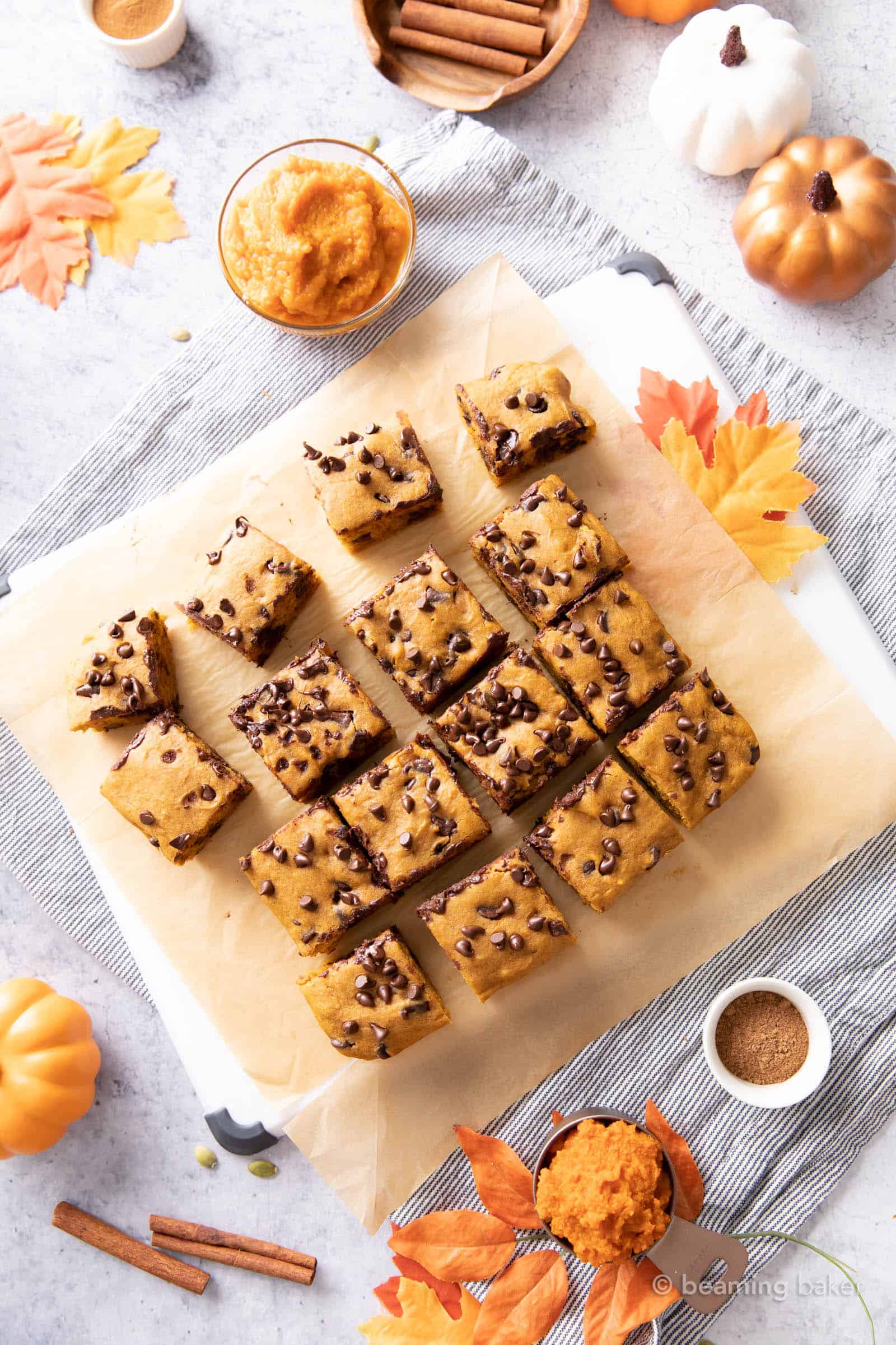 kitchen table with sliced pumpkin chocolate chip bars on parchment paper with fall leaves, pumpkin, and spices