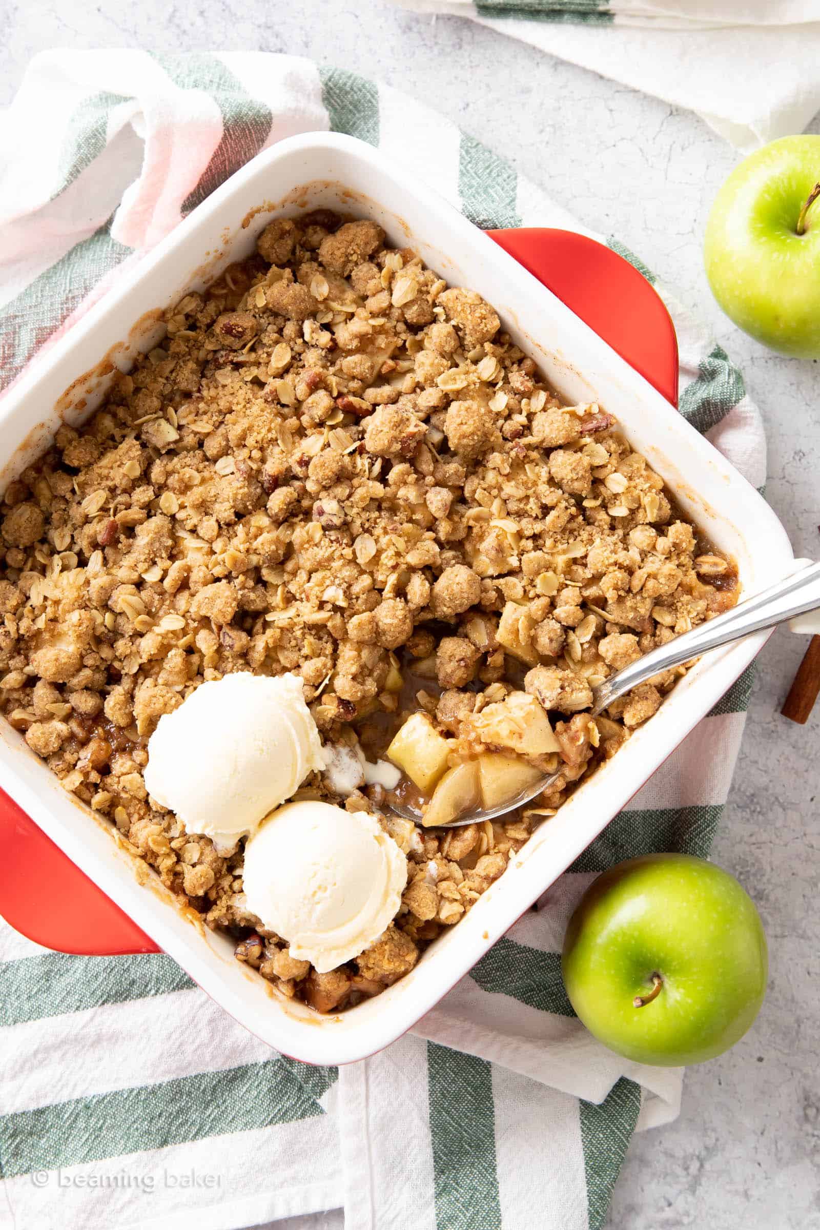 Two scoops of vanilla ice cream on top of a pan of vegan apple crumble with a green apple on the side