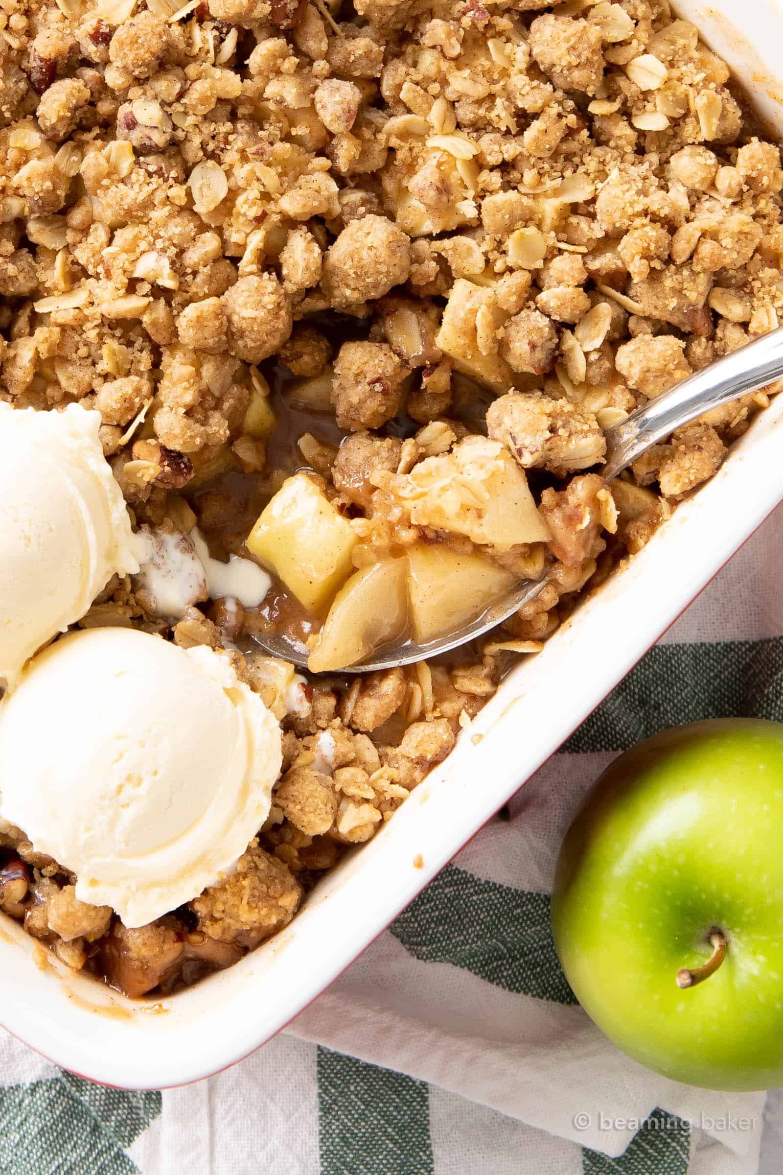 A silver spoon scooping up apple crumble from a baking dish with ice cream