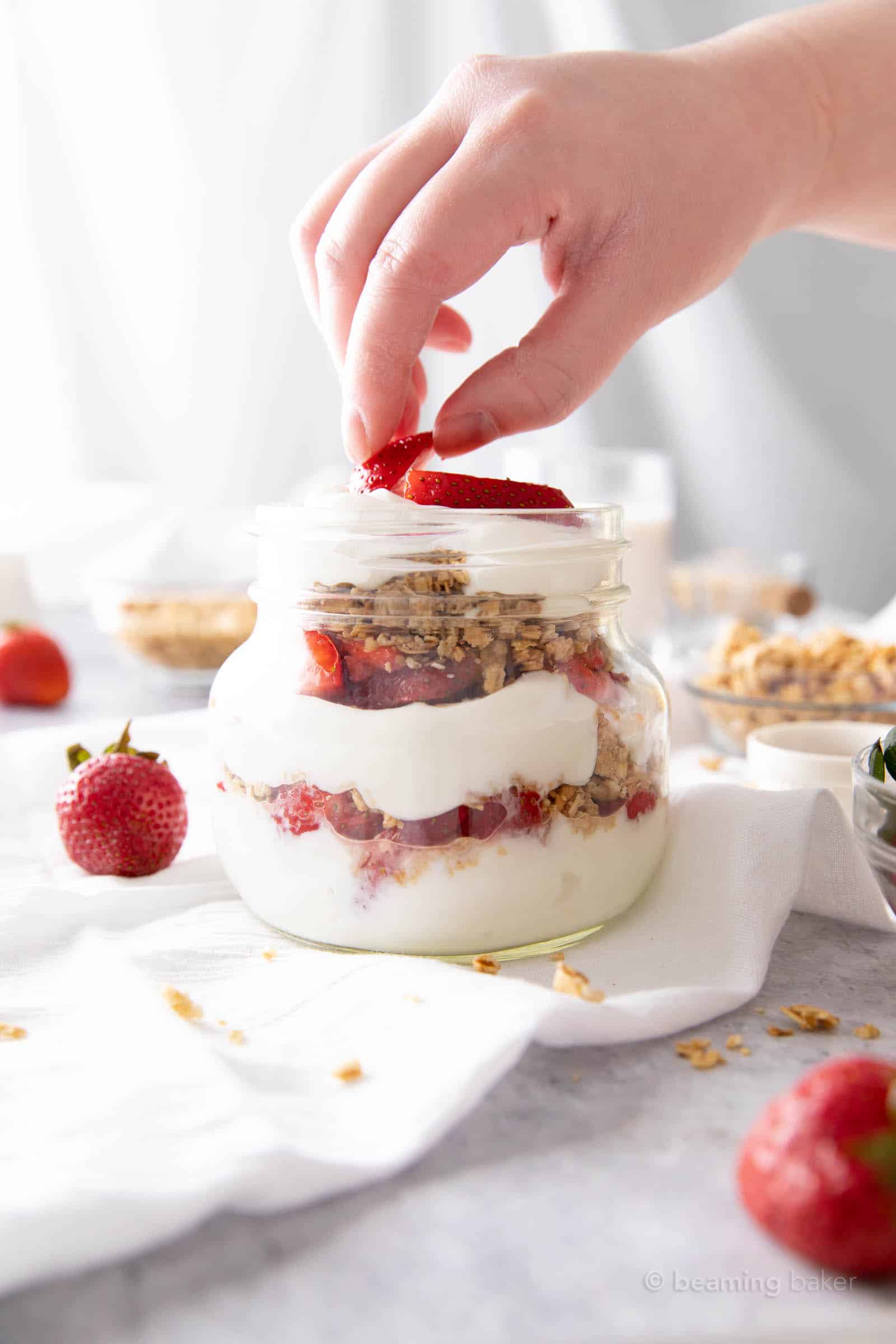 Hand placing a second strawberry slice atop a layer of yogurt in the parfait