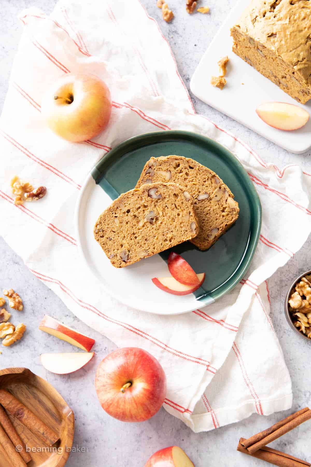 two slices of apple cinnamon bread with walnuts on a green and white plate atop a cloth with apples