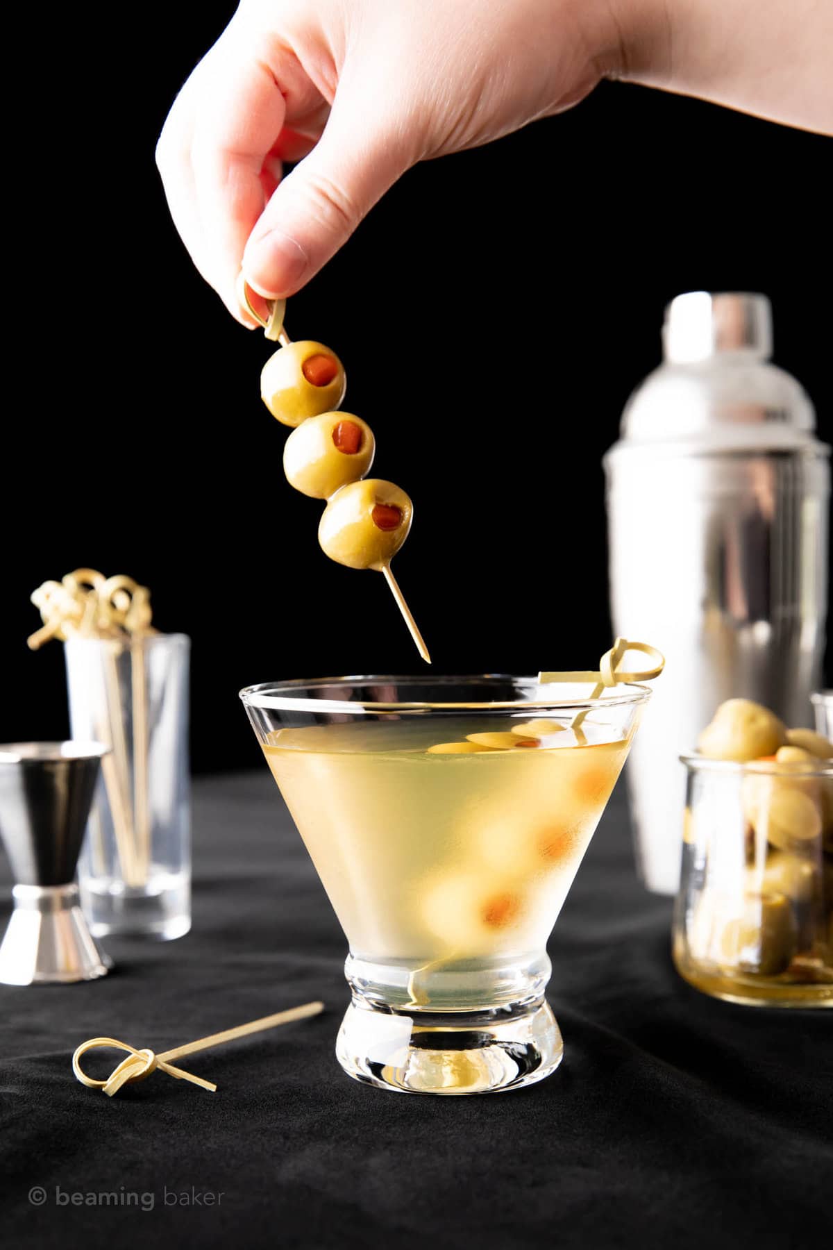 Hand holding a second olive skewer to complete the extra dirty martini against a black backdrop