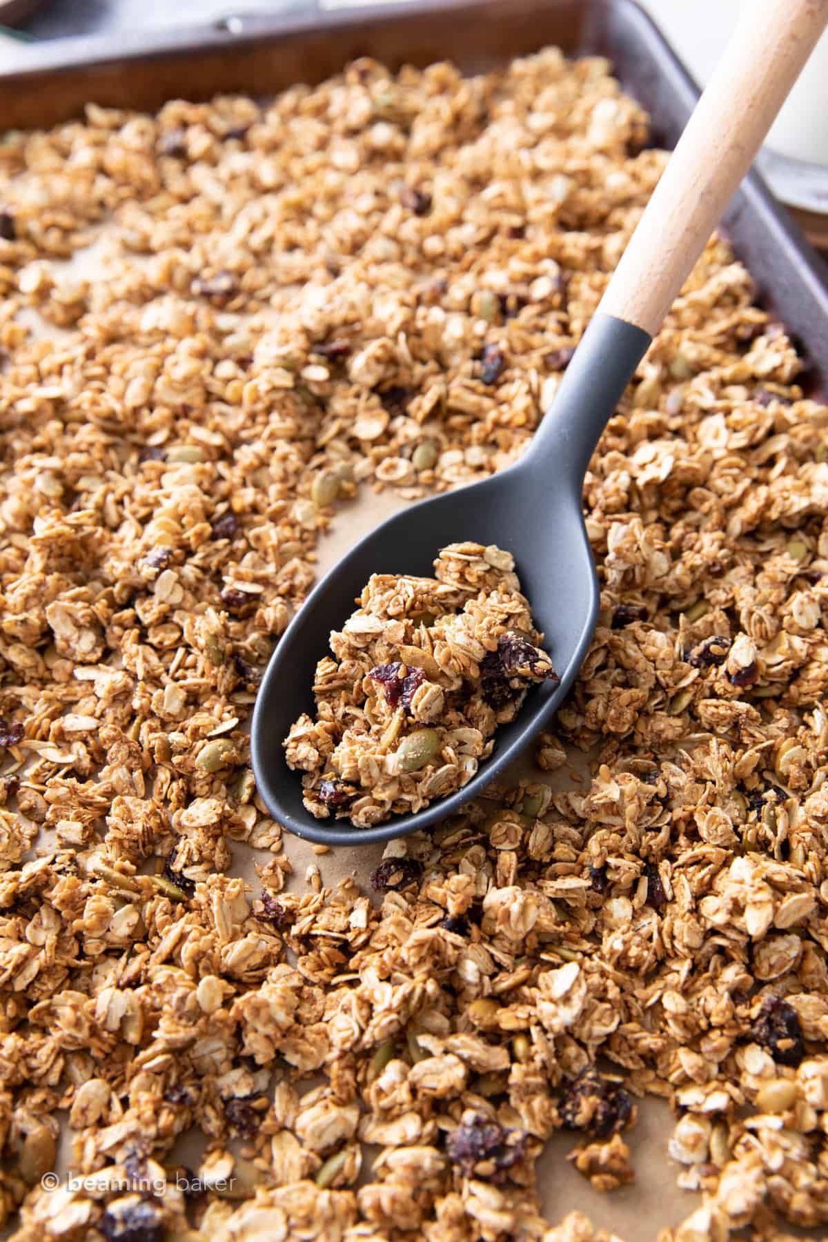 Large gray spoon filled with granola with flax seed sitting on a baking sheet filled with granola