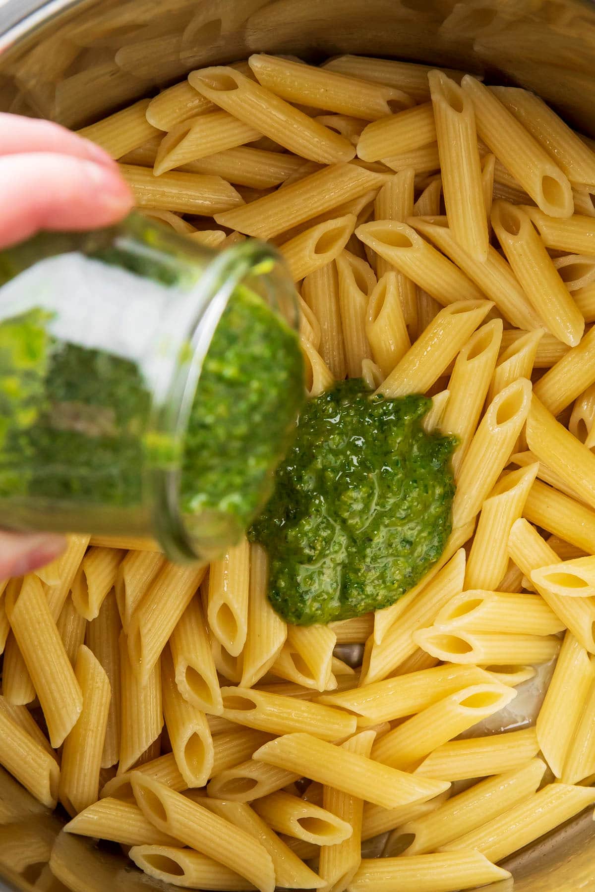 Someone pouring a jar of store bought pesto over penne pasta before stirring together to make pesto pasta with pine nuts.