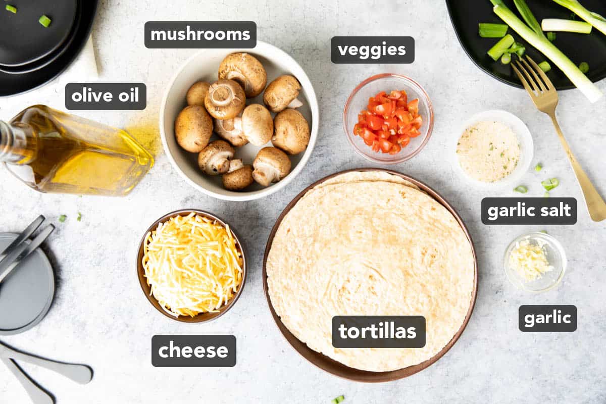 Mushroom quesadilla ingredients including mushrooms, veggies, tortillas, and cheese laid out on a kitchen table