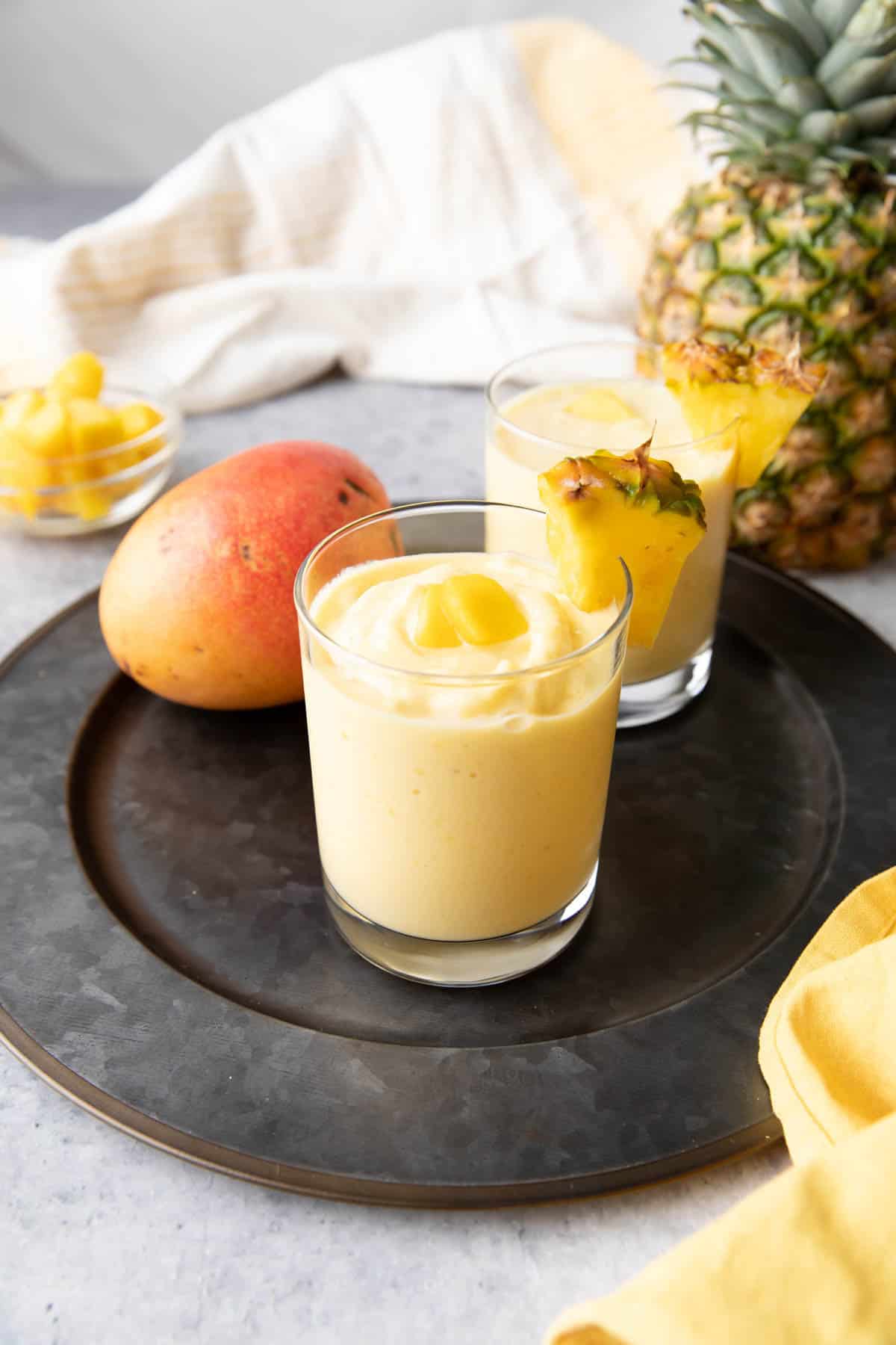a kitchen table with a mango, a pineapple, and two Mango Pineapple Smoothies