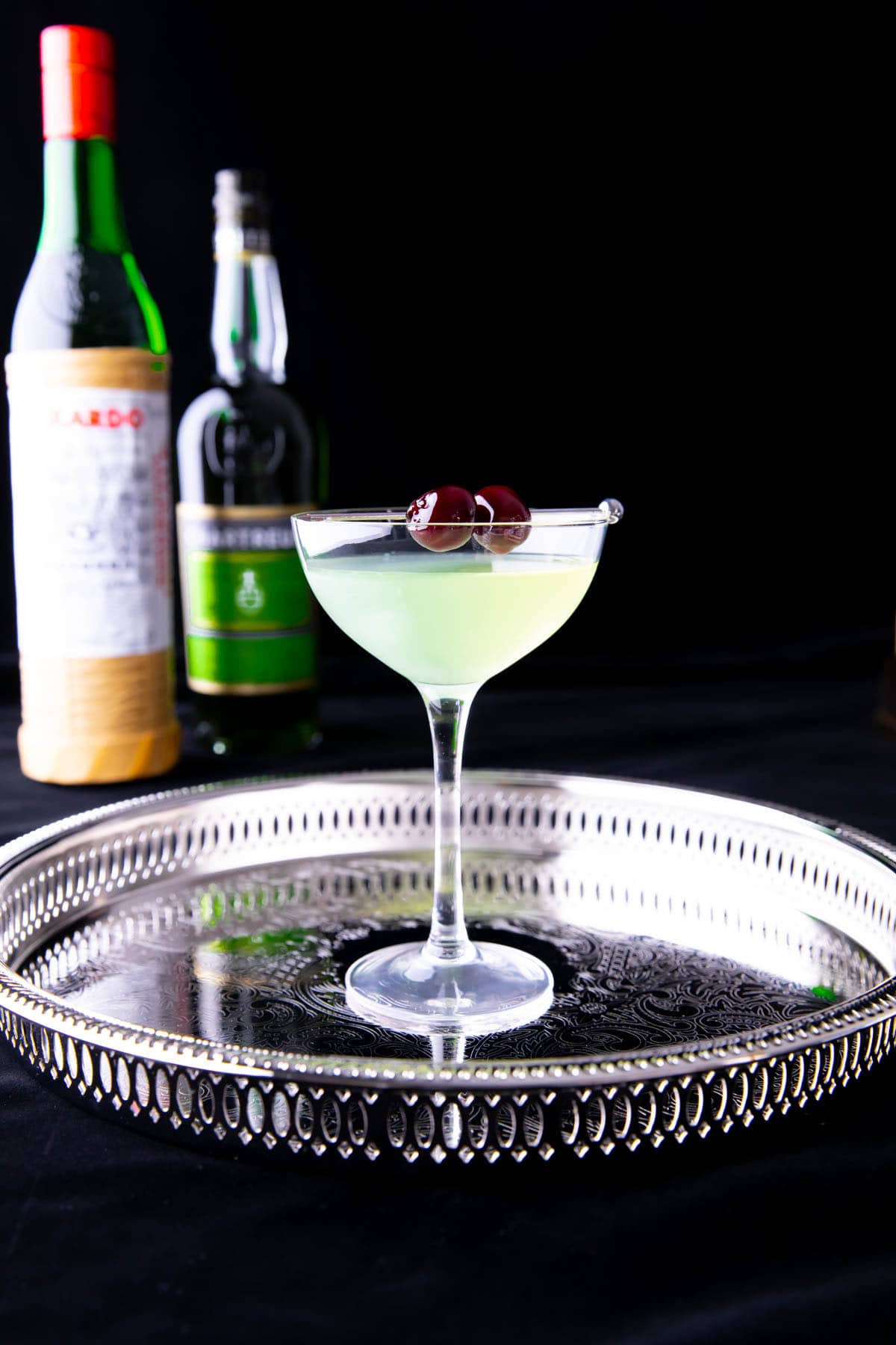 A bottle of Luxardo maraschino liqueur and Green Chartreuse with a serving tray of this cocktail