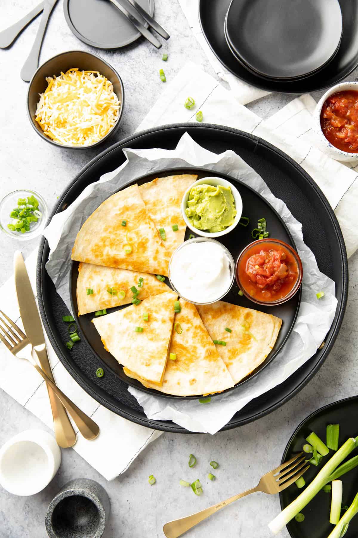 dining table with green onion-garnished Mexican dish served with tomato salsa, guacamole, and sour cream with utensils