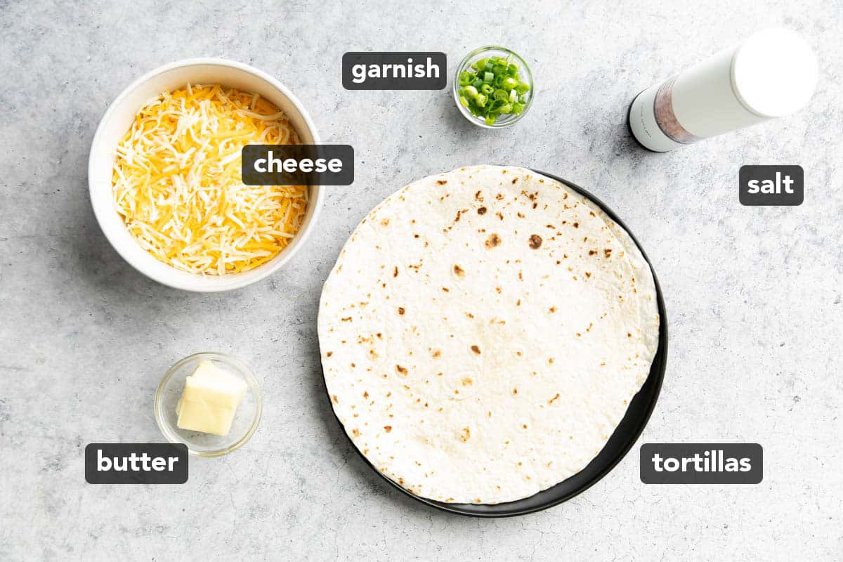 Shredded cheese, butter, flour tortillas, and salt laid out on a kitchen table ahead of preparing this recipe