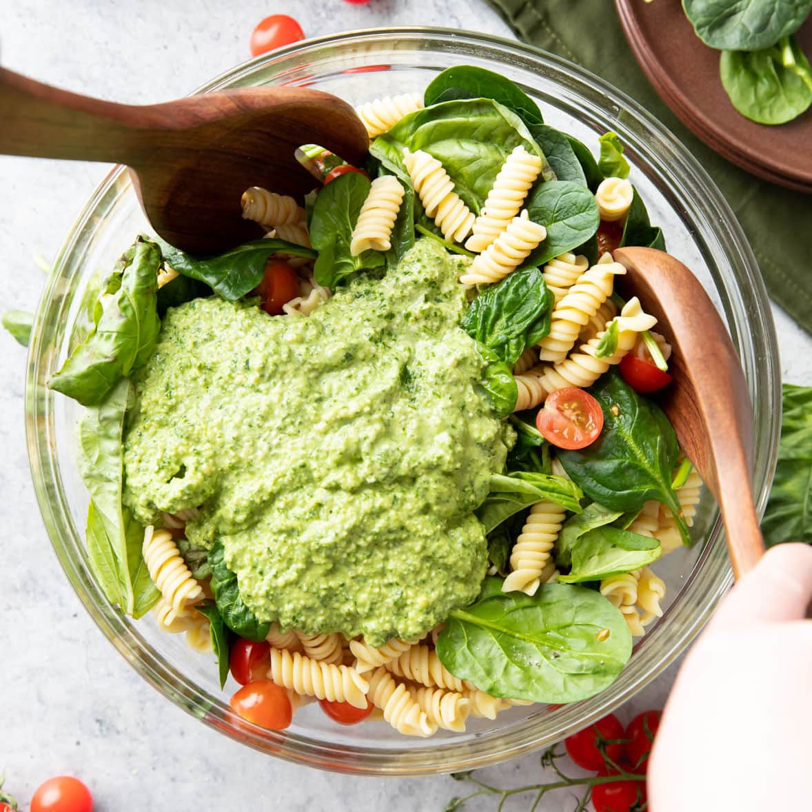 Hands holding two wooden salad tongs to mix pesto sauce onto fresh basil and tomato pasta salad
