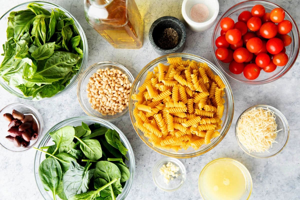 Ingredients for pasta salad with pesto and feta including spinach, basil, pasta, cheese, tomatoes, pesto sauce, olives, olive oil, salt and pepper.
