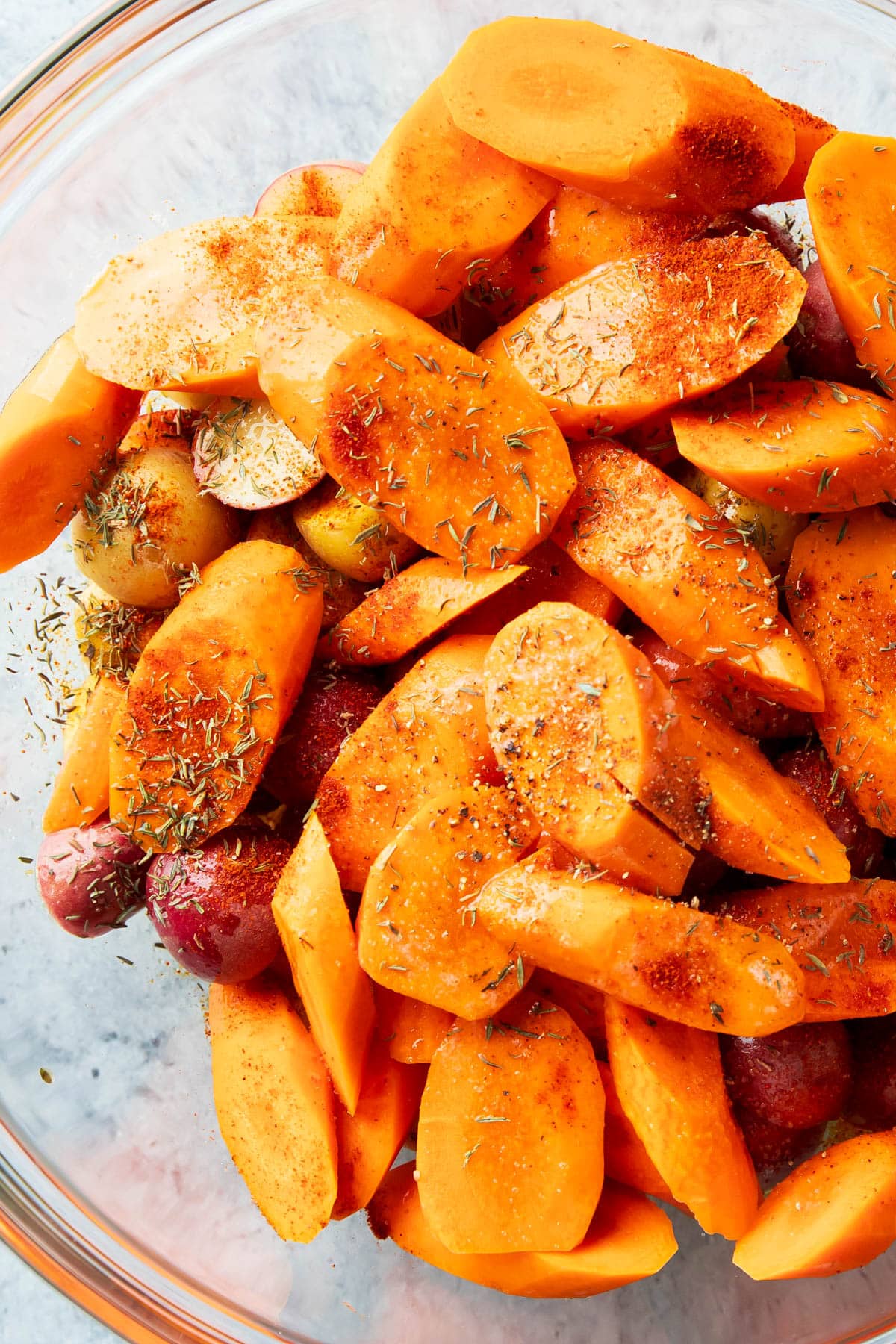 close up of thyme, paprika, salt, and more spices tossed over carrots and potatoes in a bowl before roasting