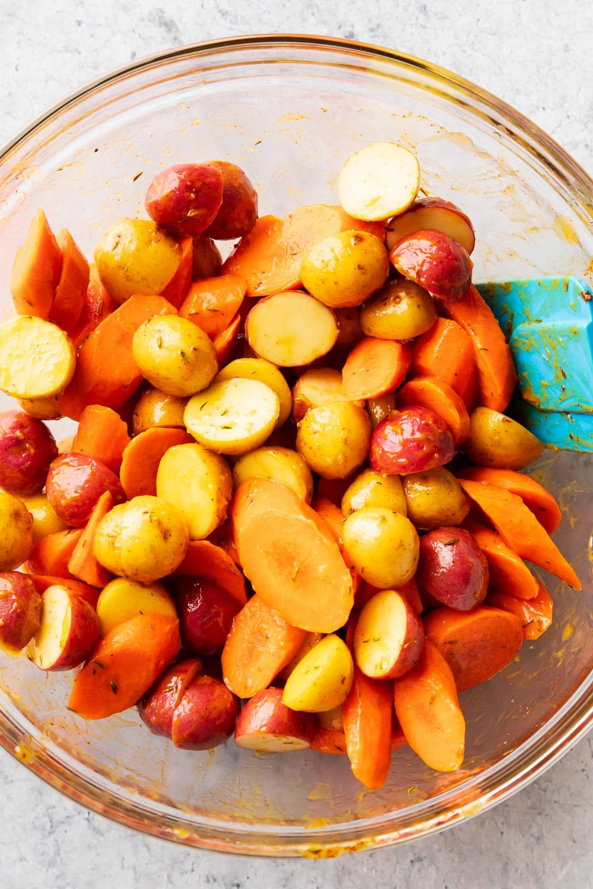 Yukon potatoes and sliced carrots tossed with butter, herbs, and spice in a glass mixing bowl with a spatula inserted