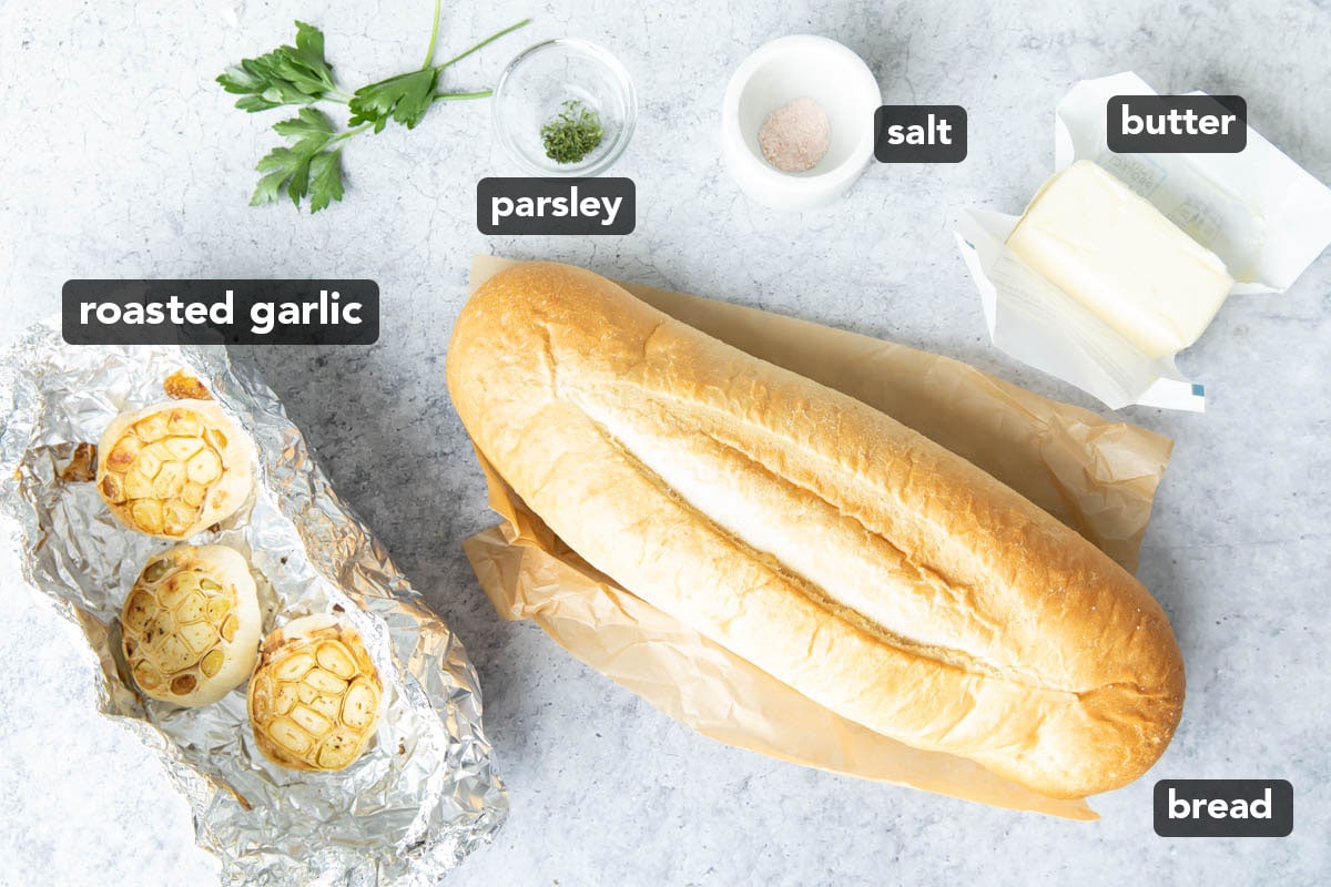 Roasted garlic bread ingredients including a loaf of bread, butter, salt, roasted garlic, and parsley on a kitchen table