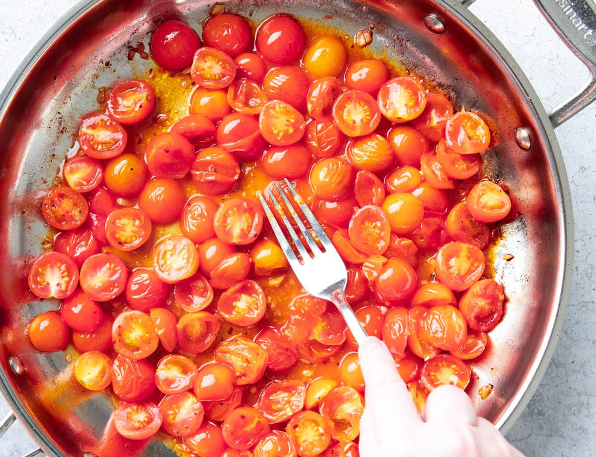 Hand using a fork to smash tomatoes into the sauce cooking on a skillet