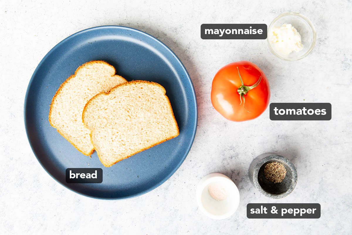Tomato sandwich ingredients on a kitchen table, including a beefsteak tomato, slices of sandwich bread, mayo, and salt and pepper