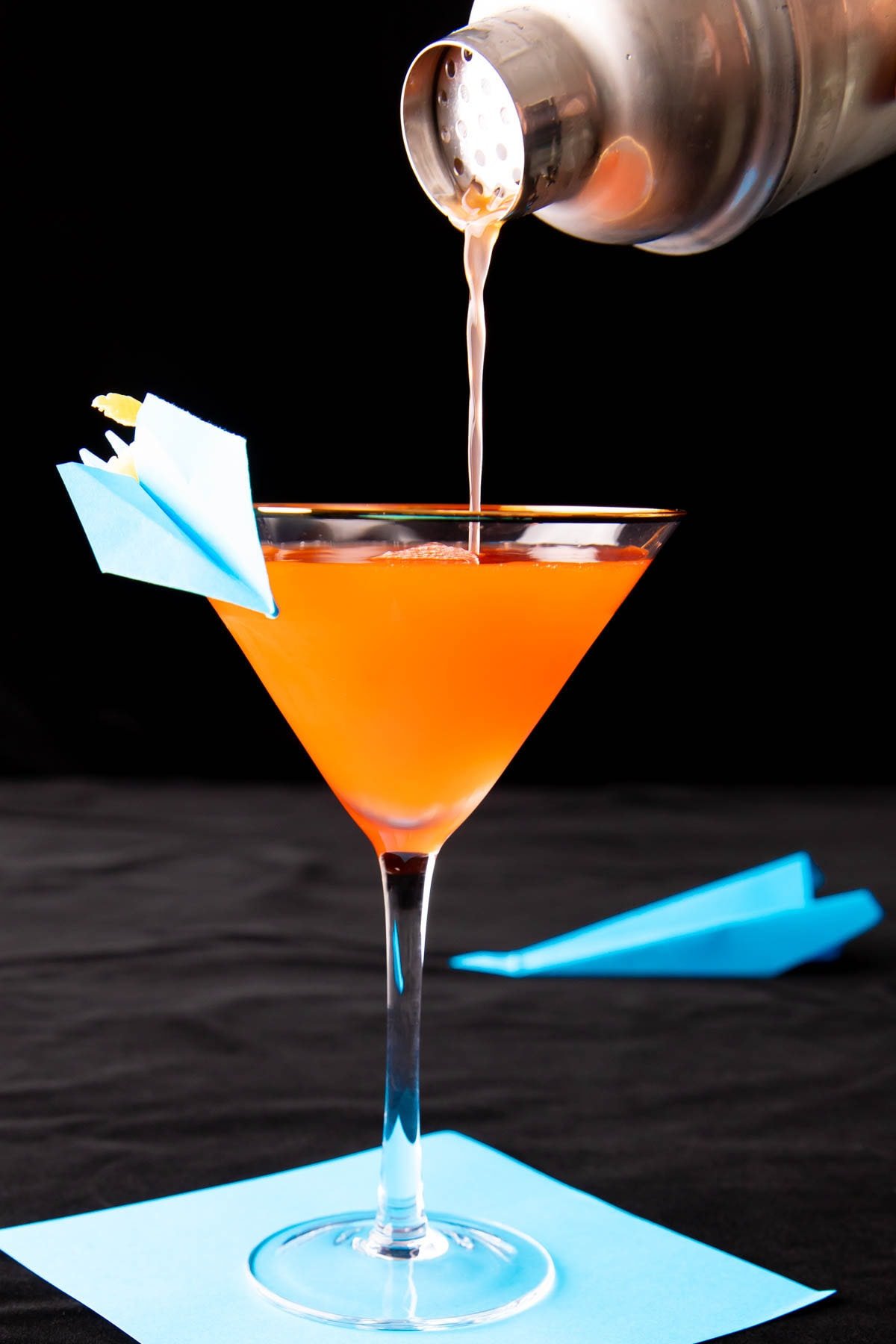 A photo showing How to Make a Paper Plane Cocktail – pouring the last drop of drink mixture into a glass with a paper plane garnish