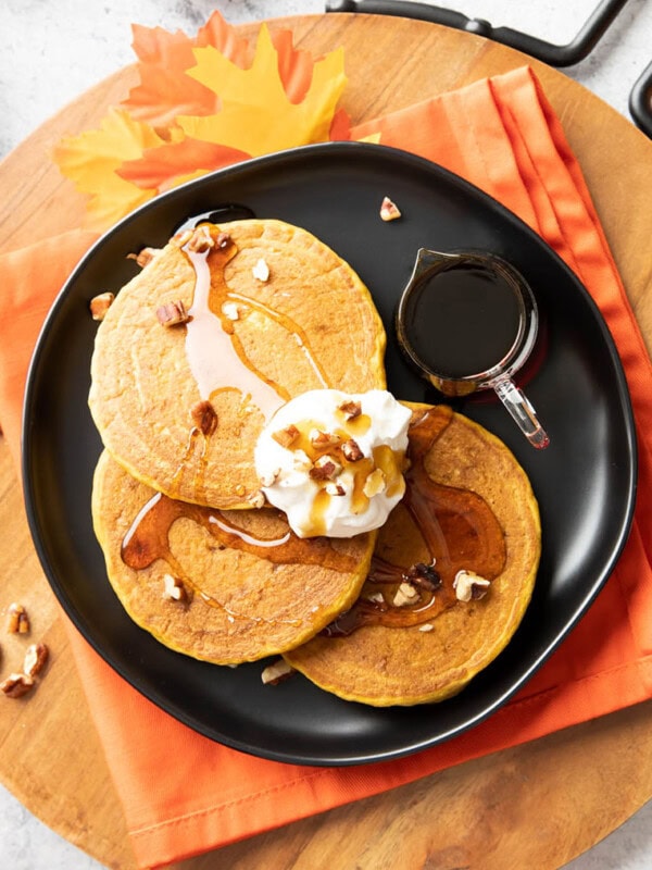 A plate full of pumpkin pancakes covered in syrup, whipped topping, and nuts